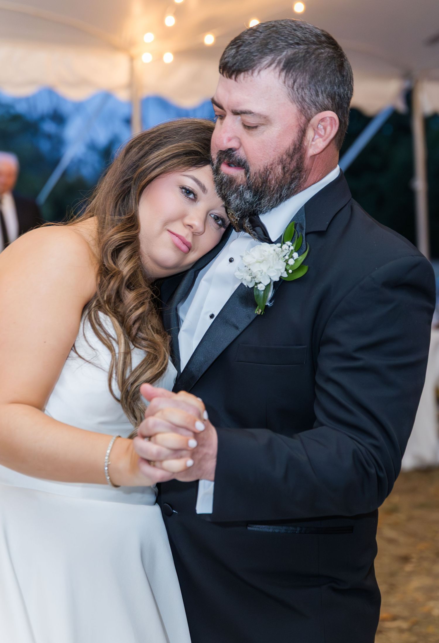 Bride and groom share an emotional first dance, with groom in black tuxedo and white boutonniere at wedding reception.