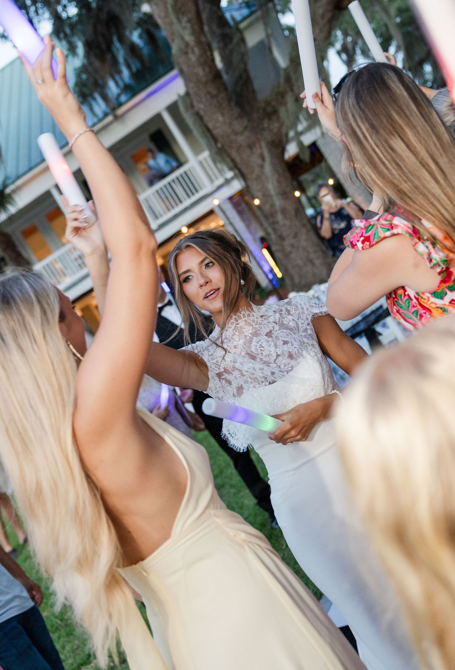 Bride in lace wedding dress dancing outdoors with guests holding colorful glow sticks at reception.