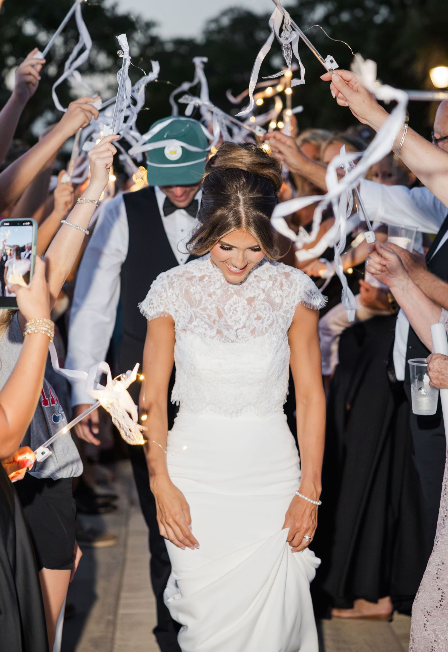 Bride in white lace gown walks through celebratory ribbon wand exit tunnel at outdoor evening wedding reception.