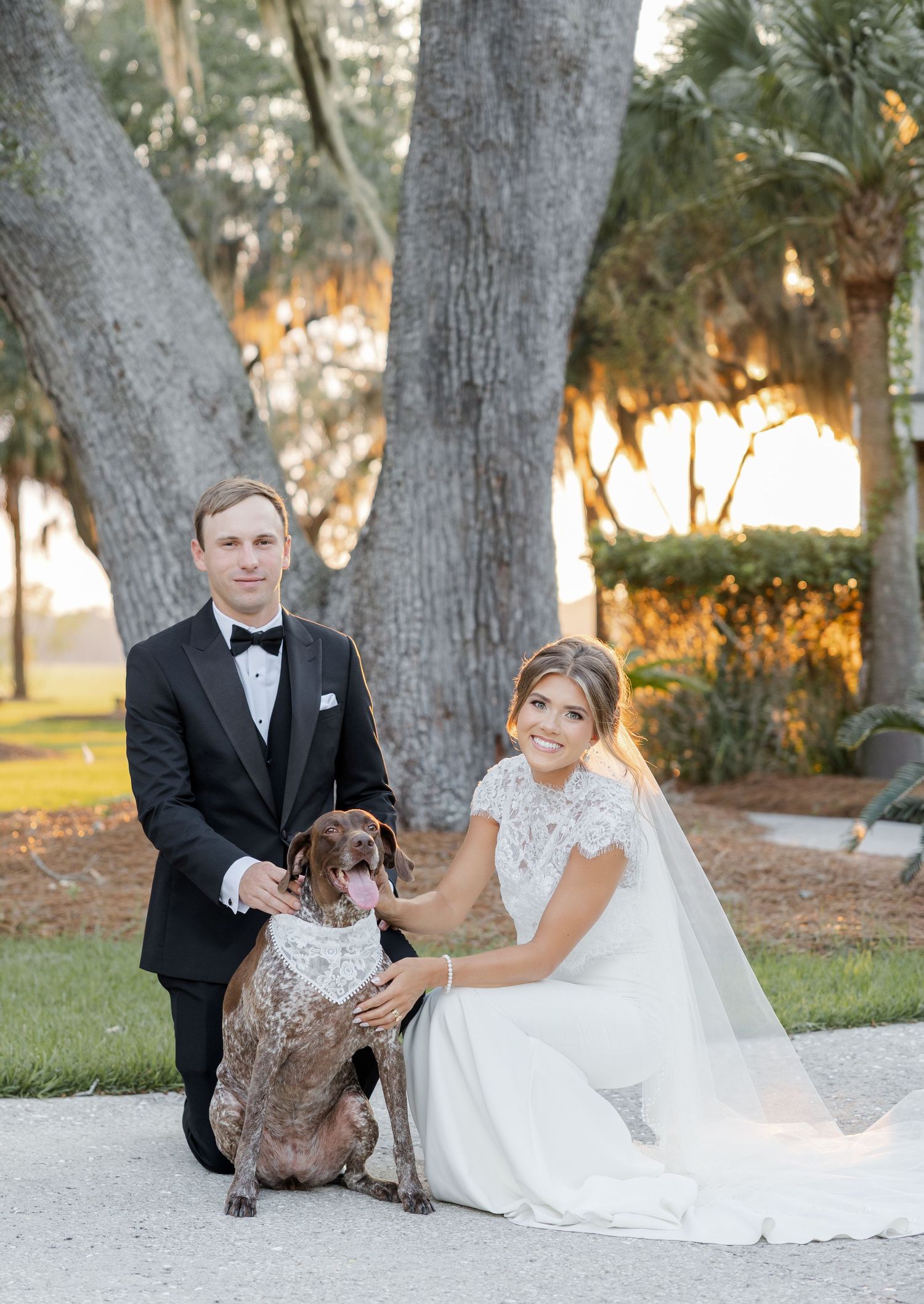 Bride and groom pose with their brown spotted dog wearing a white collar outdoors near large oak trees at golden hour.