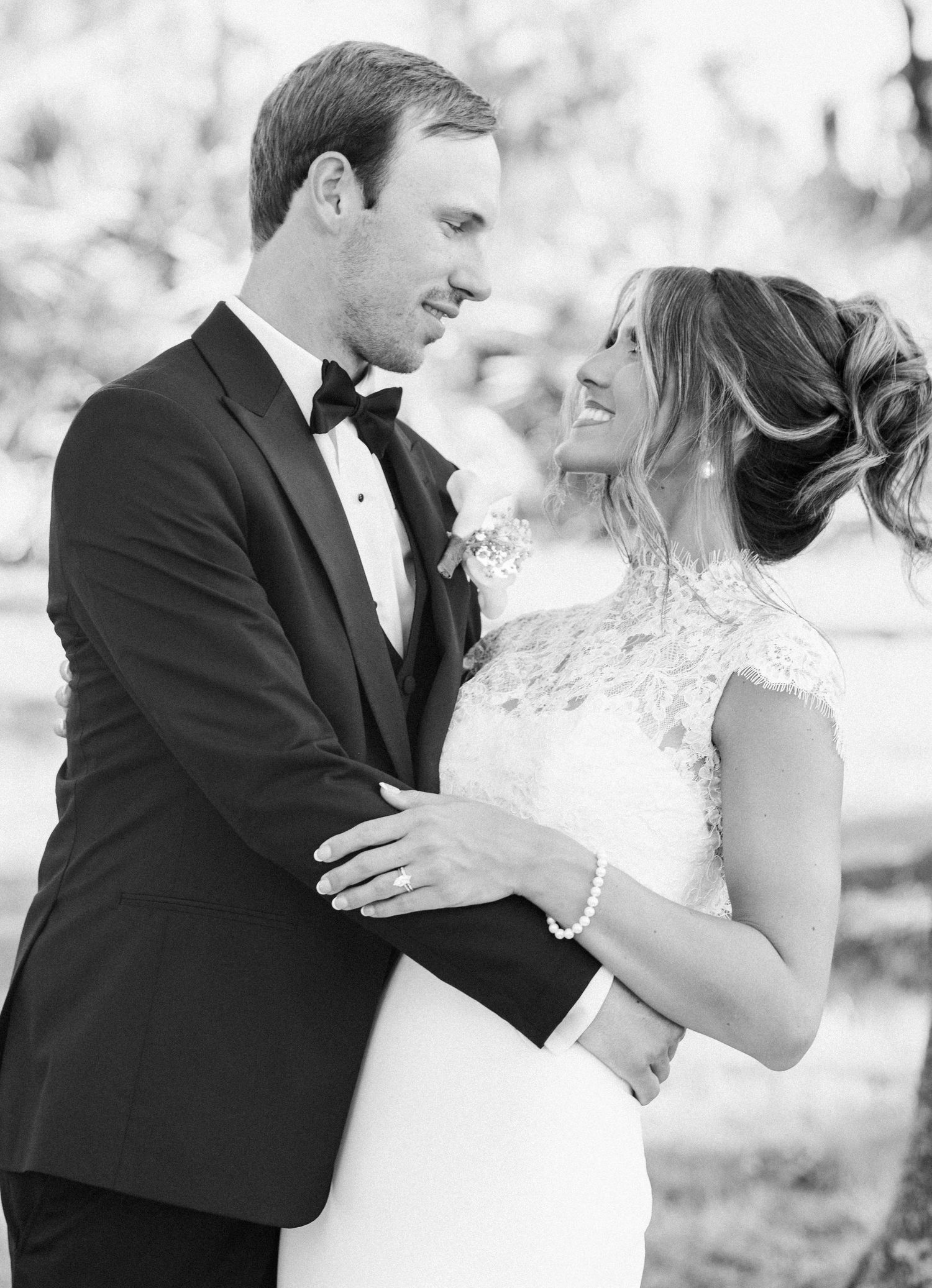 Black and white wedding photo of smiling bride in lace gown and groom in tuxedo embracing outdoors.