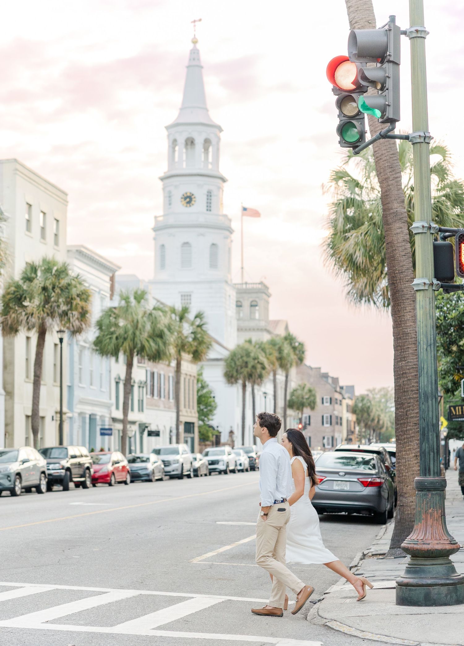 Couple crossing a historic Charleston street with a white church steeple and palm trees in the background.