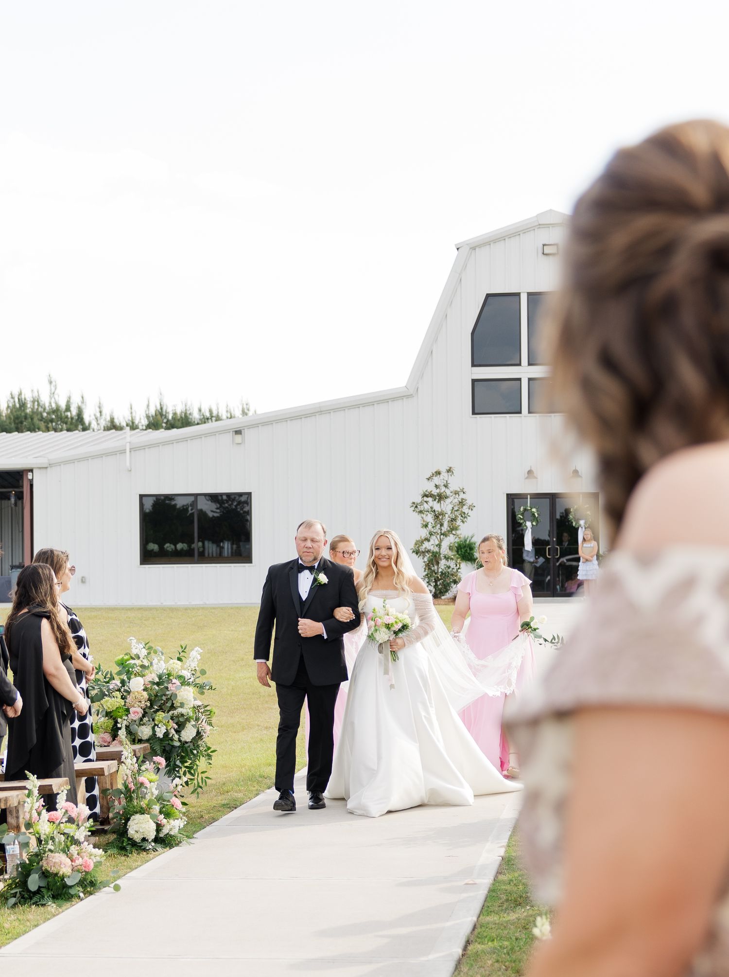 Bride in white gown walks down outdoor aisle with escort toward white barn venue.