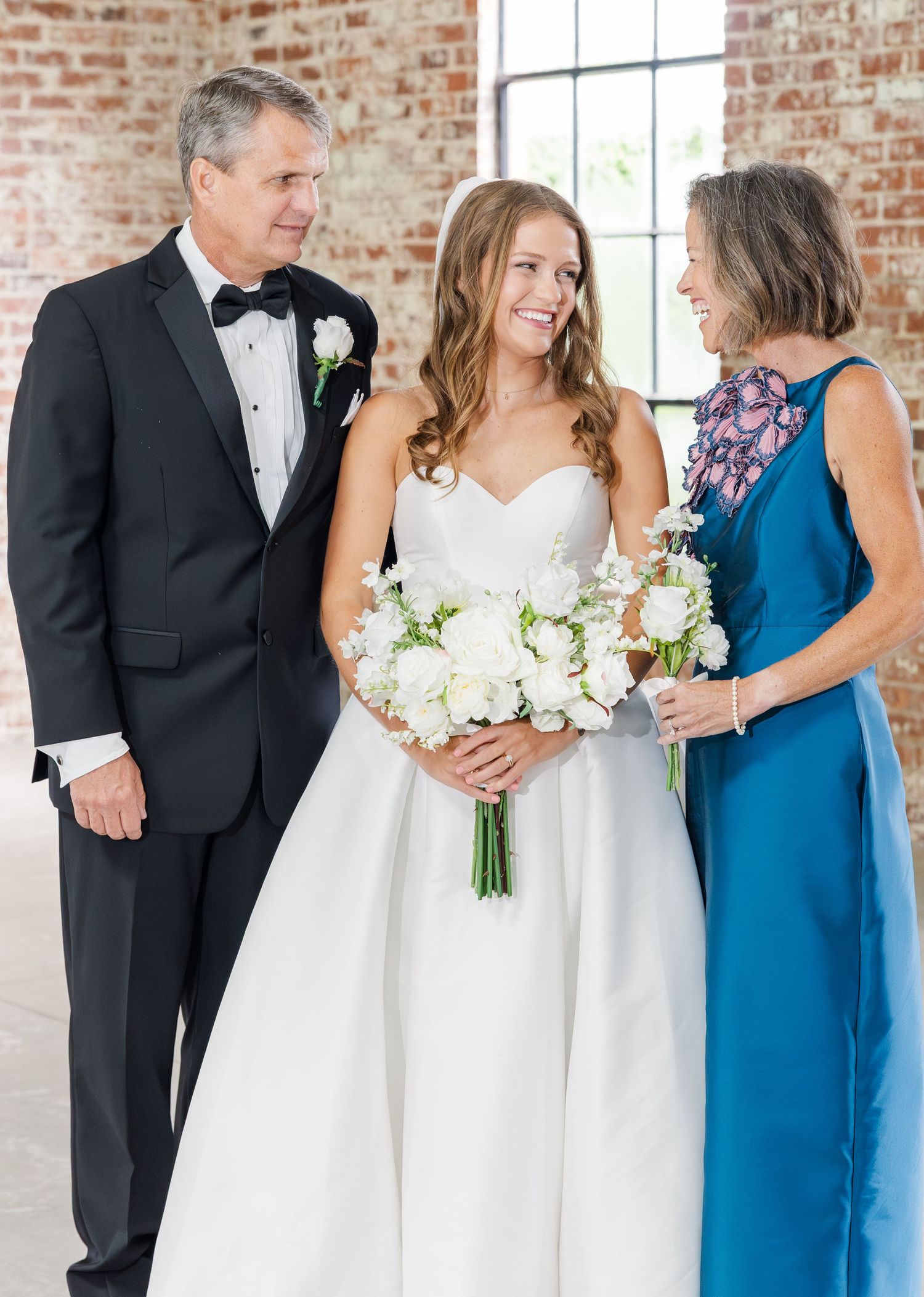 Bride holding white bouquet smiles with parents in elegant wedding attire at brick venue.