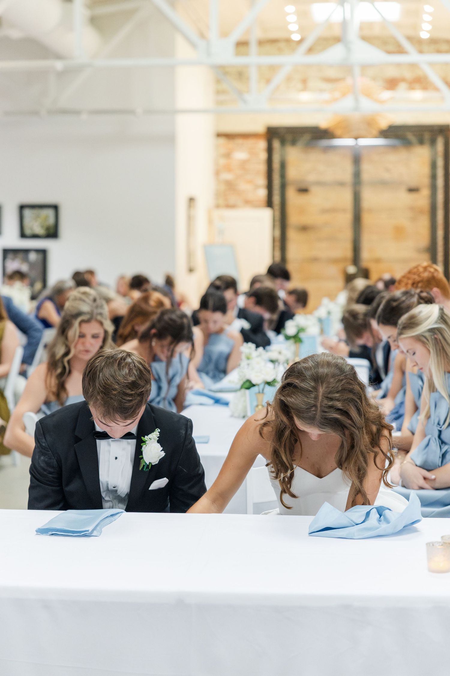 Bride and groom bow heads in prayer at sweetheart table during wedding reception.