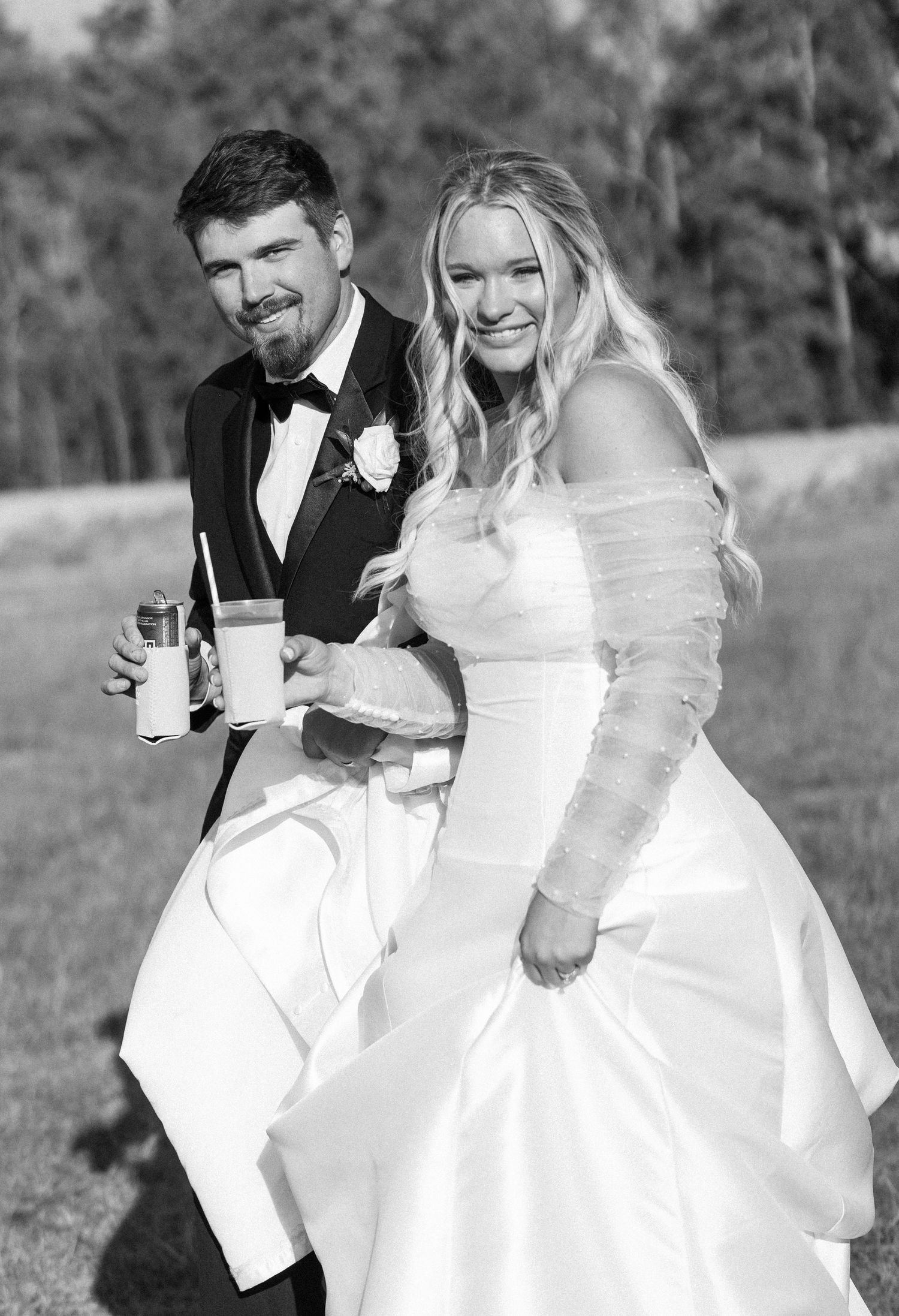 Black and white photo of smiling bride and groom holding drinks outdoors on wedding day.