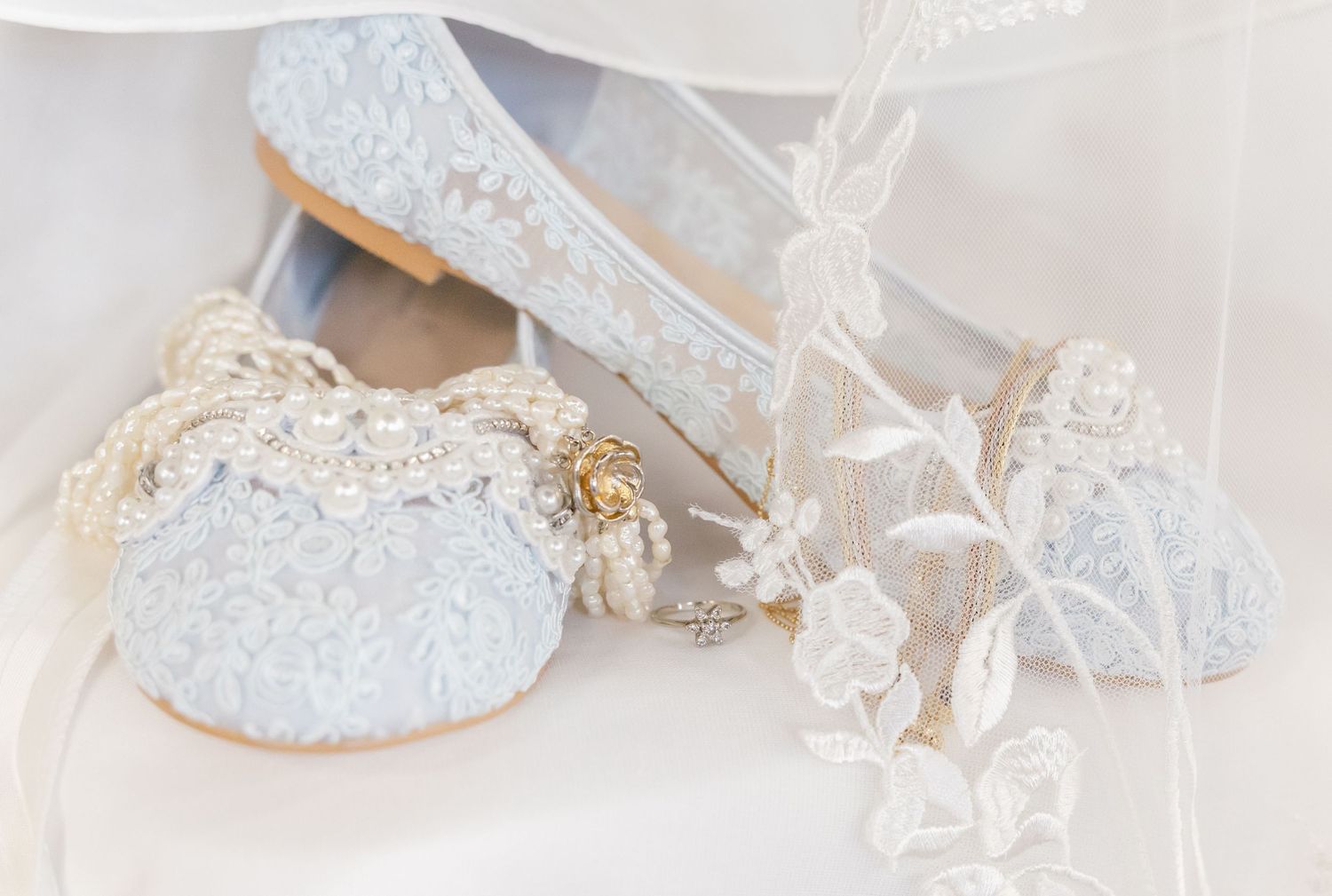 Delicate light blue lace bridal flats adorned with pearl embellishments and gold bee brooch displayed with sheer veil.
