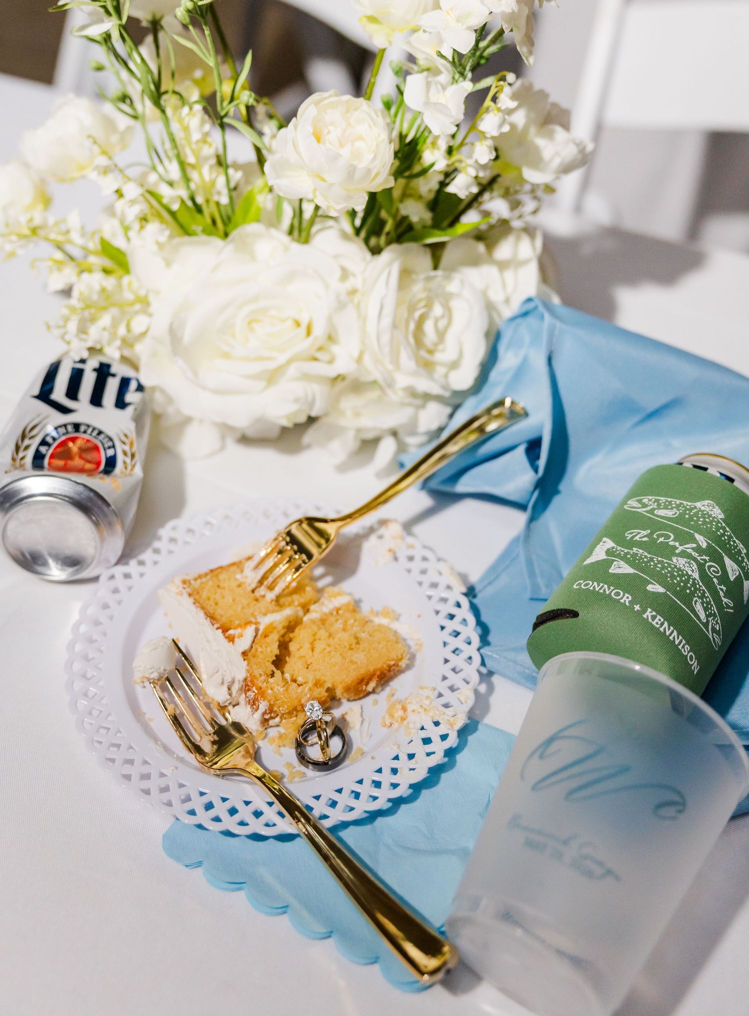 Wedding reception table setting with gold fork, cake slice, Miller Lite, white roses, blue napkin.