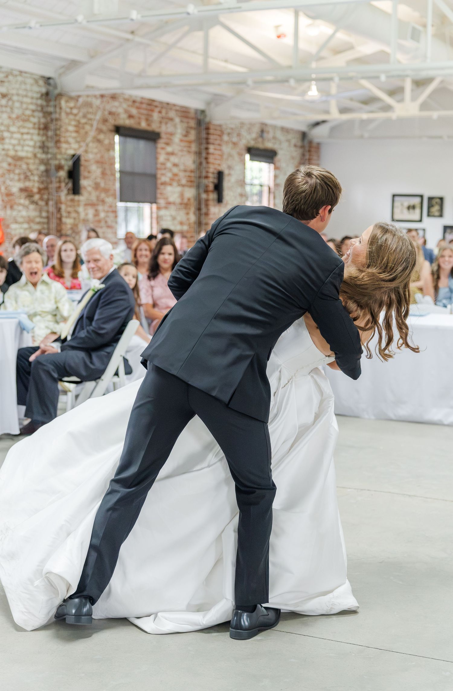 Groom dips bride during first dance at elegant wedding reception with guests watching in background.