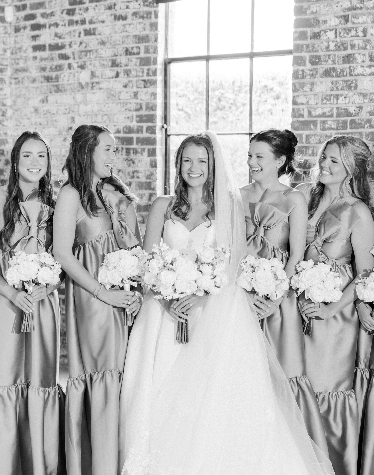 Black and white photo of bride in strapless gown with four bridesmaids in satin dresses holding white bouquets.