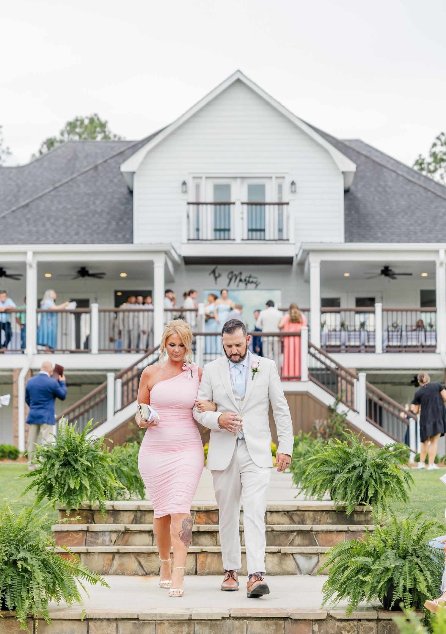 Groom in tan suit escorts woman in pink dress down steps outside elegant white wedding venue.
