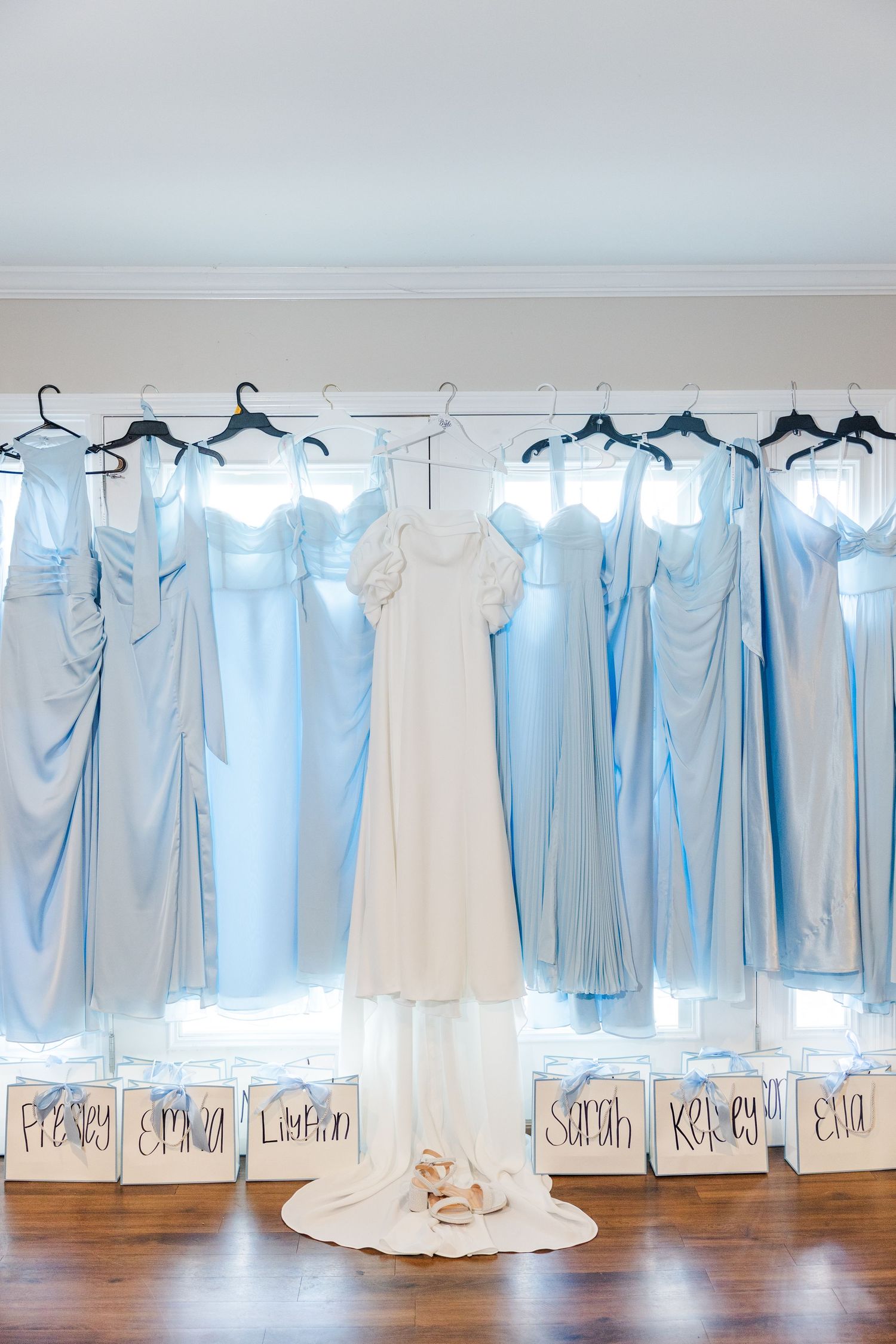 White wedding gown flanked by light blue bridesmaid dresses hanging above personalized gift boxes.