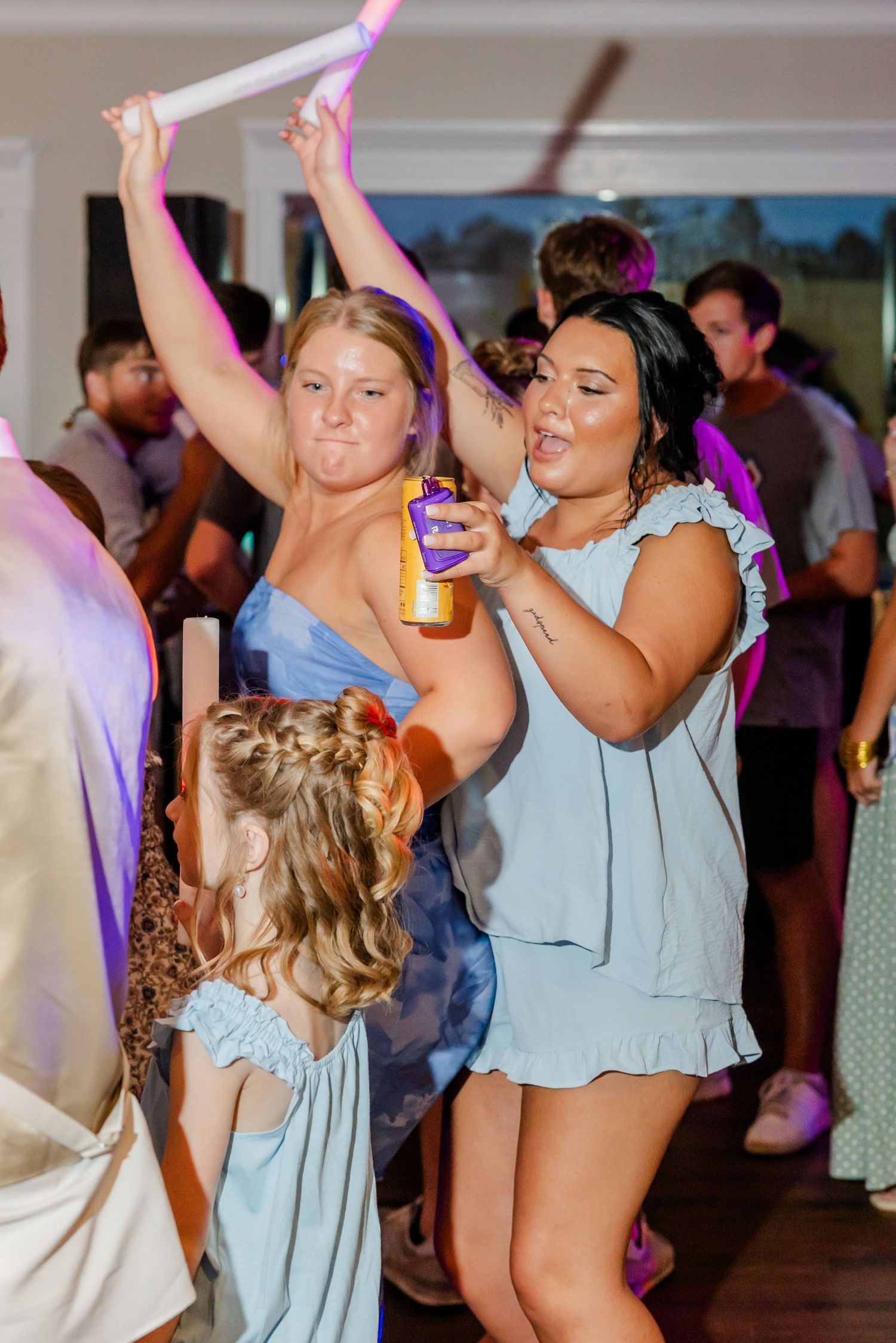 Guests in blue dresses dance energetically with glow sticks and drinks at lively wedding reception.
