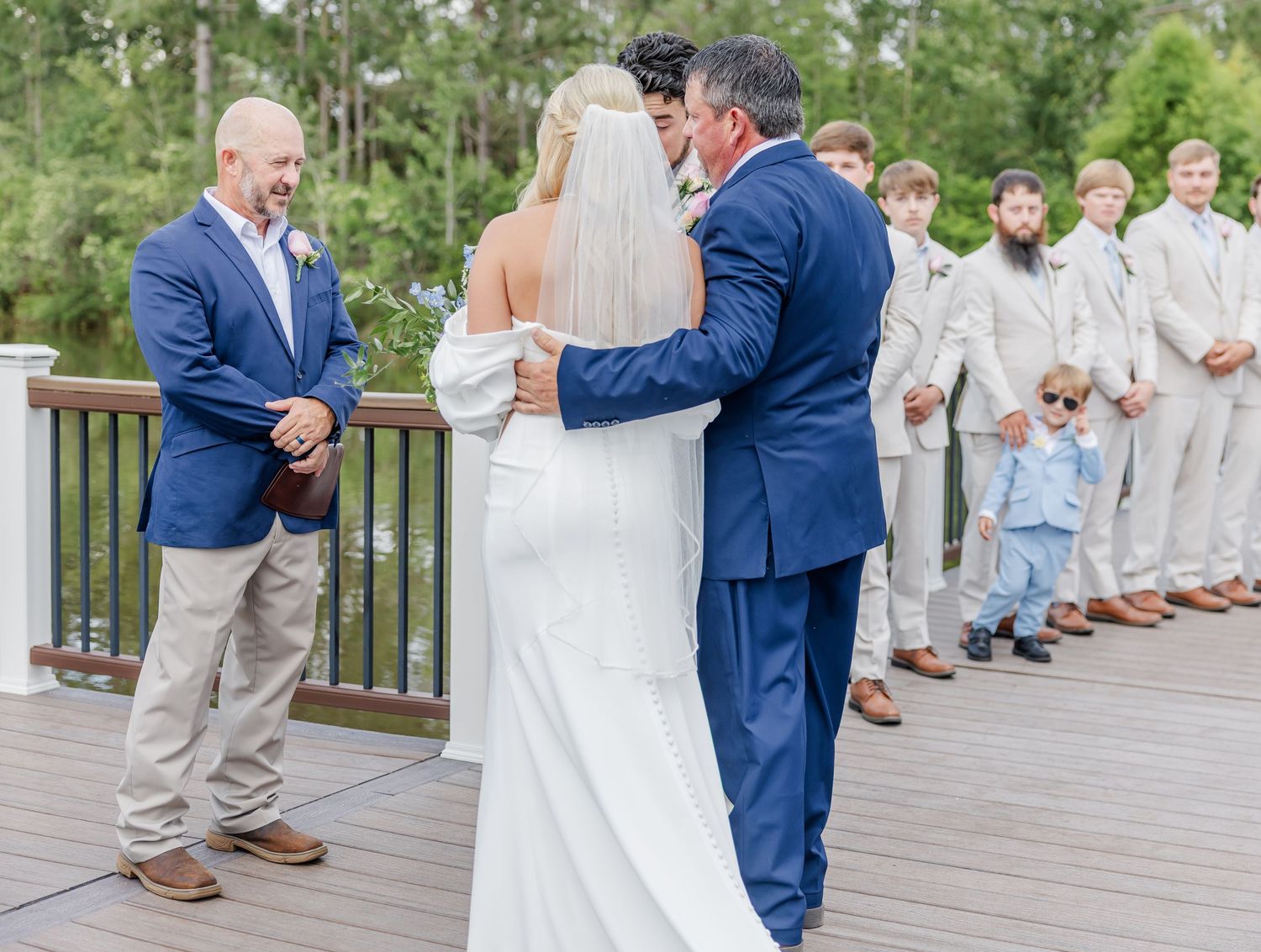 Father walks bride down aisle toward officiant and groomsmen on outdoor wooden deck ceremony.