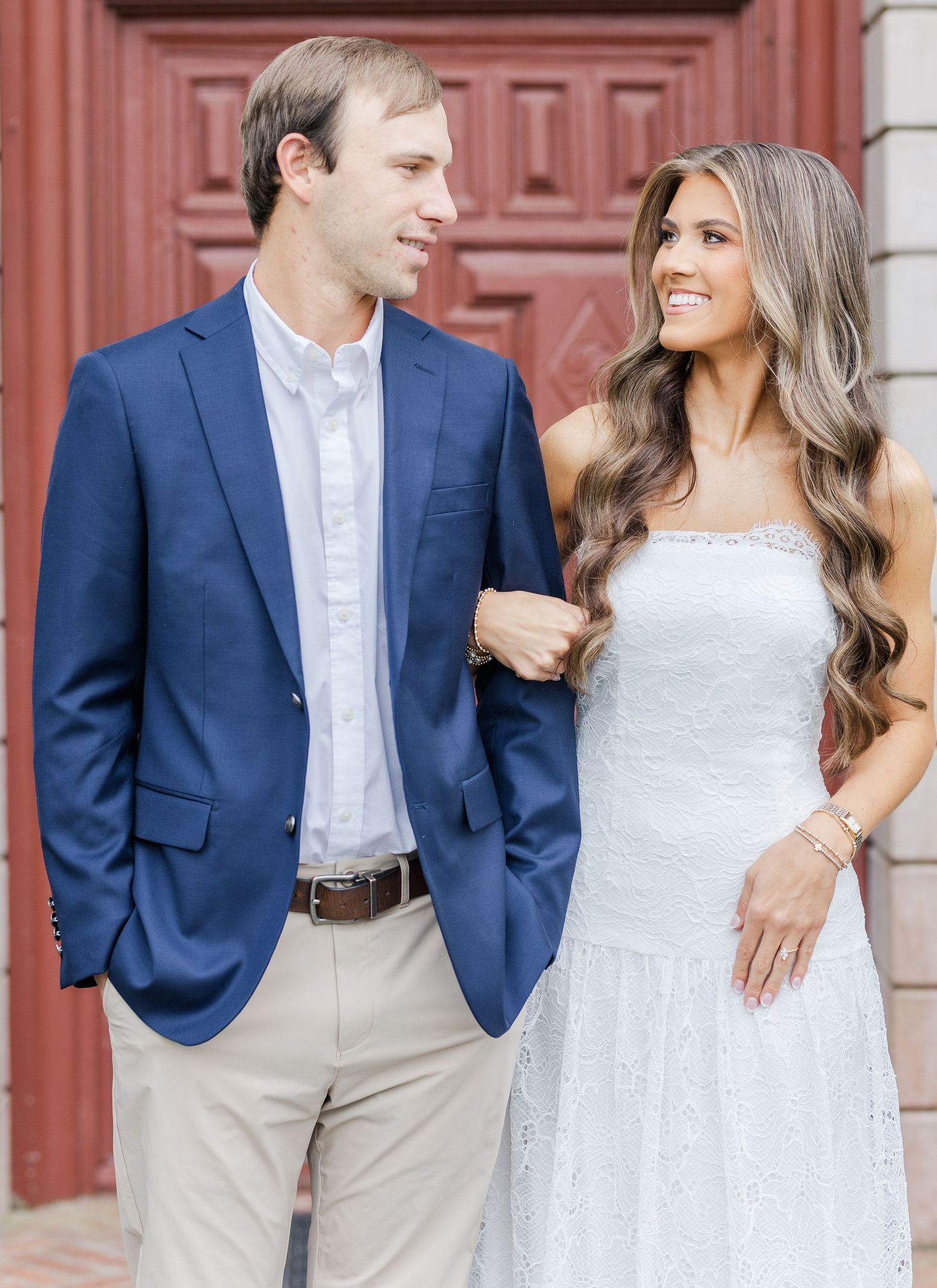 Couple in elegant engagement photo, man in navy blazer and woman in white lace dress smiling outdoors.