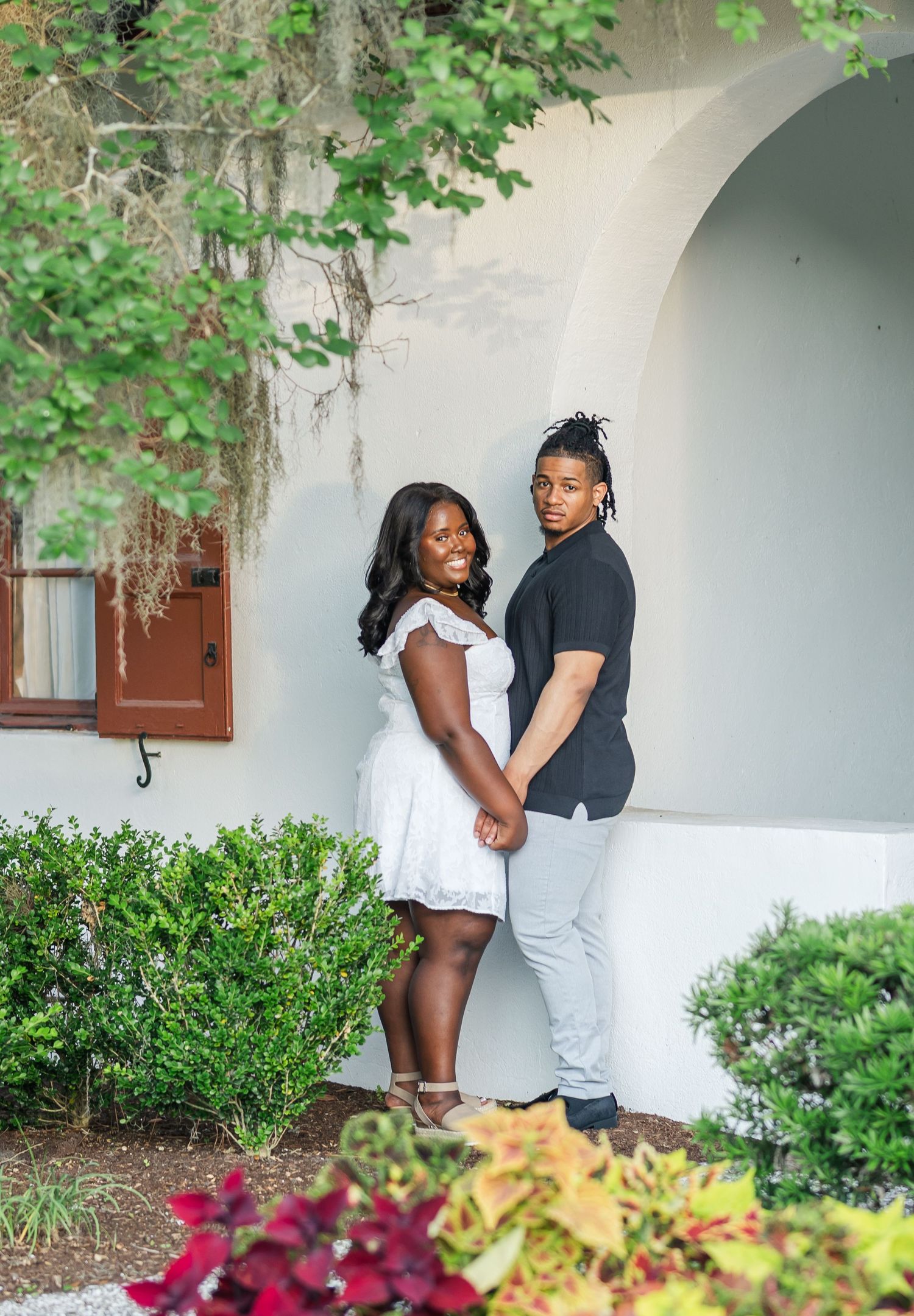 Couple holding hands and posing under a white archway surrounded by lush green landscaping.