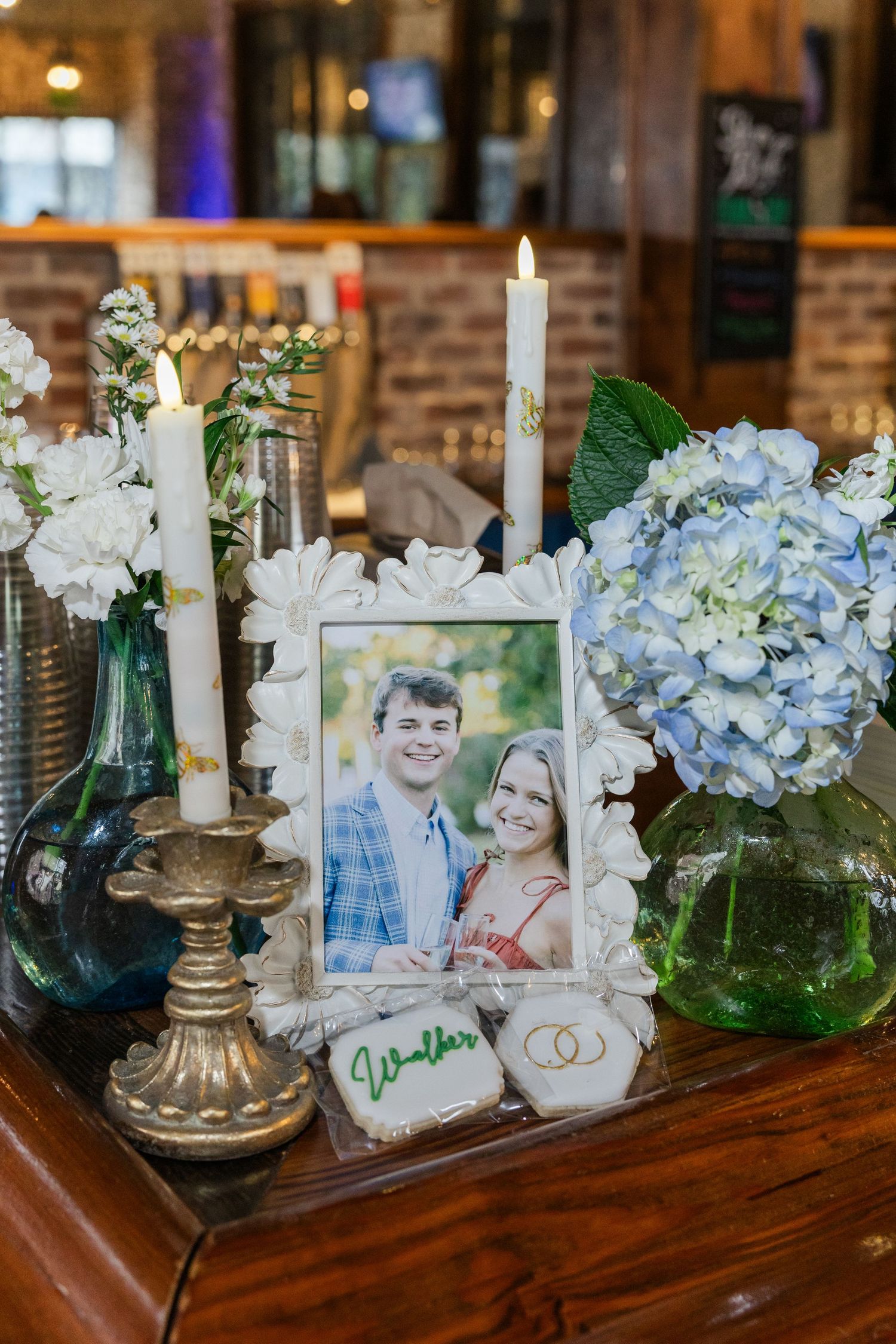 Wedding display with couple's photo in white frame, candles, blue hydrangeas, and decorated cookies on wooden table.