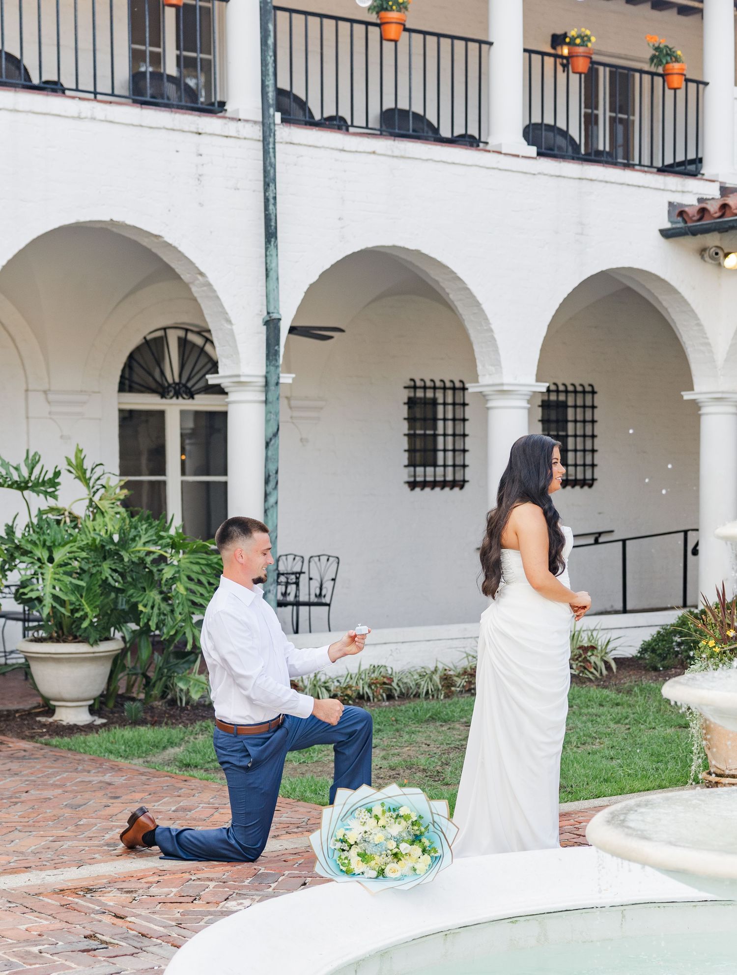Man proposes on one knee to woman in white dress by fountain at elegant white arched building.