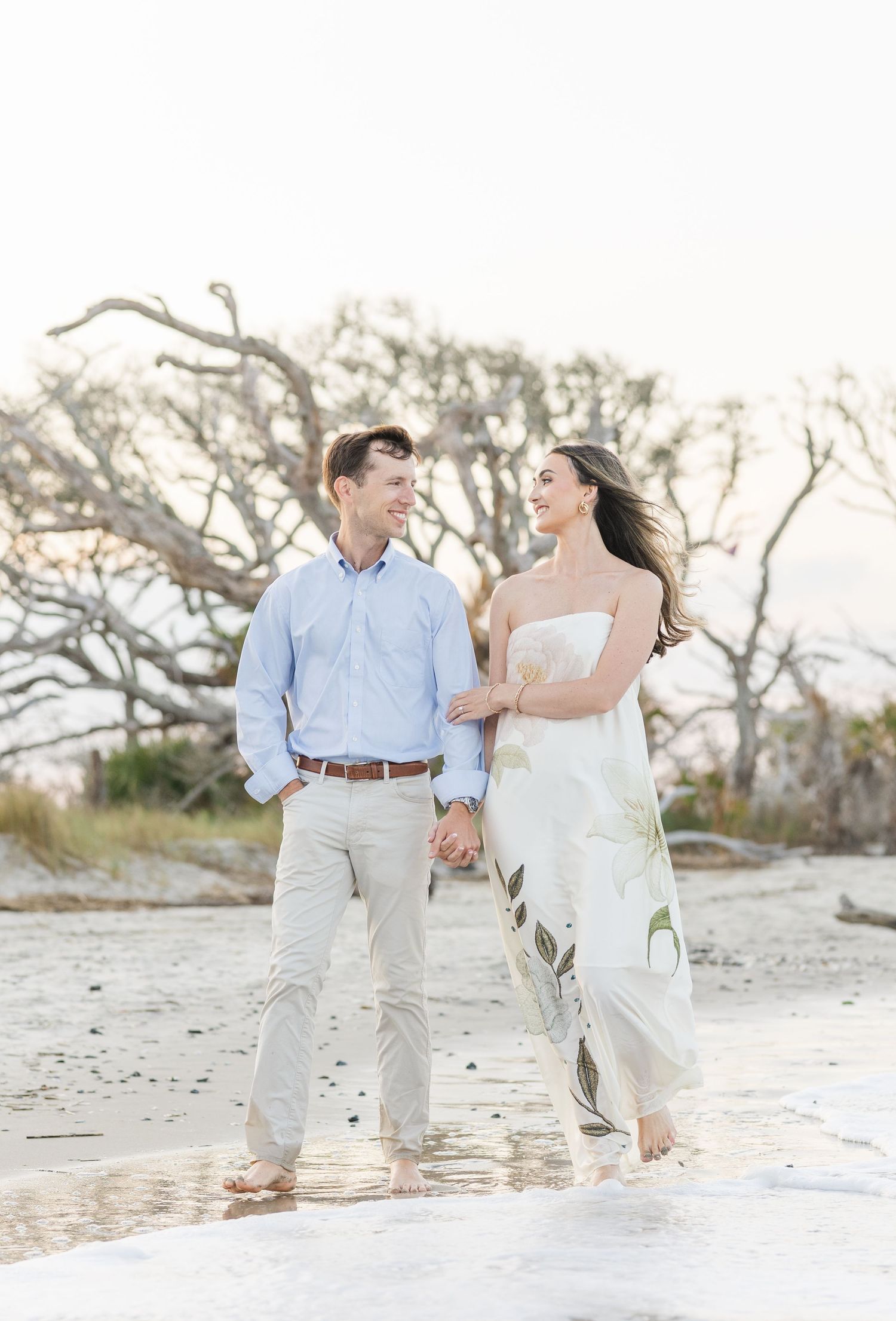Happy couple walking hand in hand on beach, woman in white dress, man in light blue shirt.