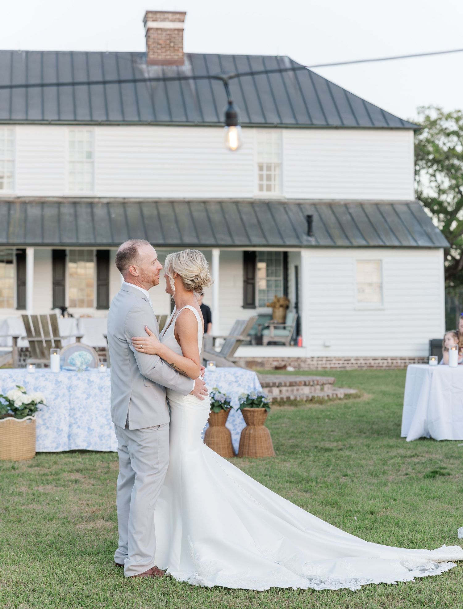 Bride and groom sharing a first dance on the lawn of a white farmhouse at dusk.