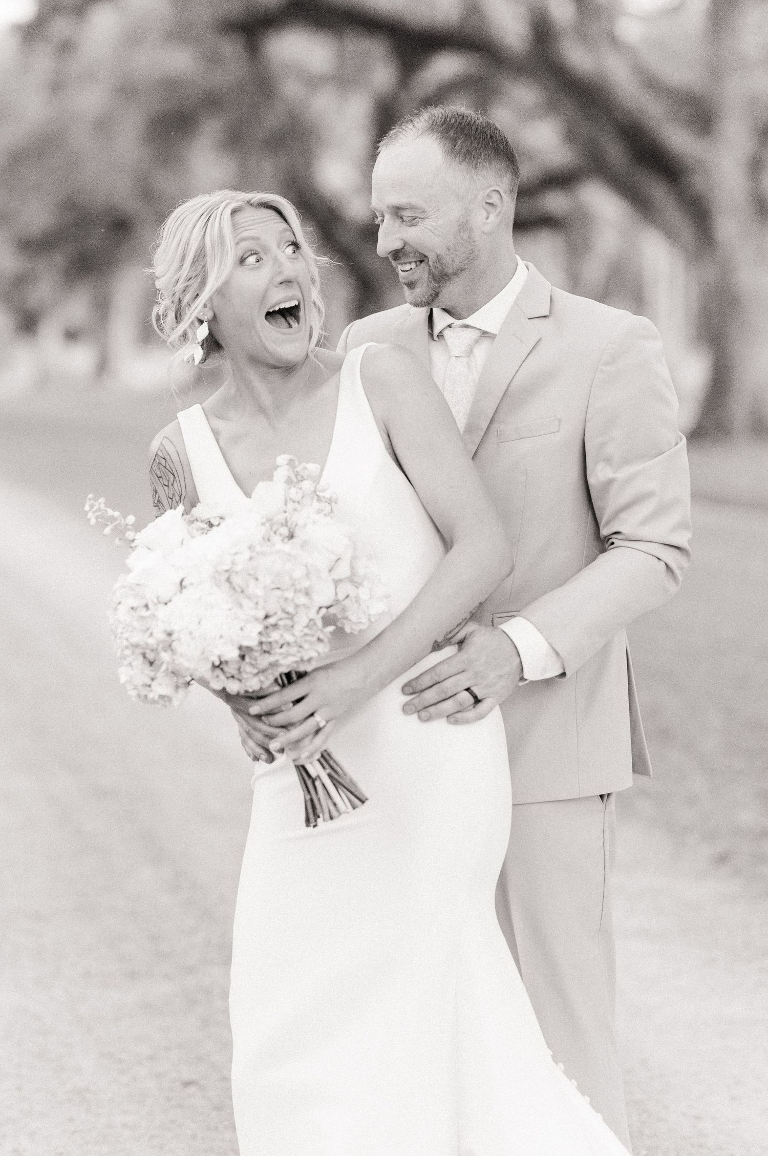 Black and white photo of laughing bride and groom embracing outdoors under large oak trees on wedding day.