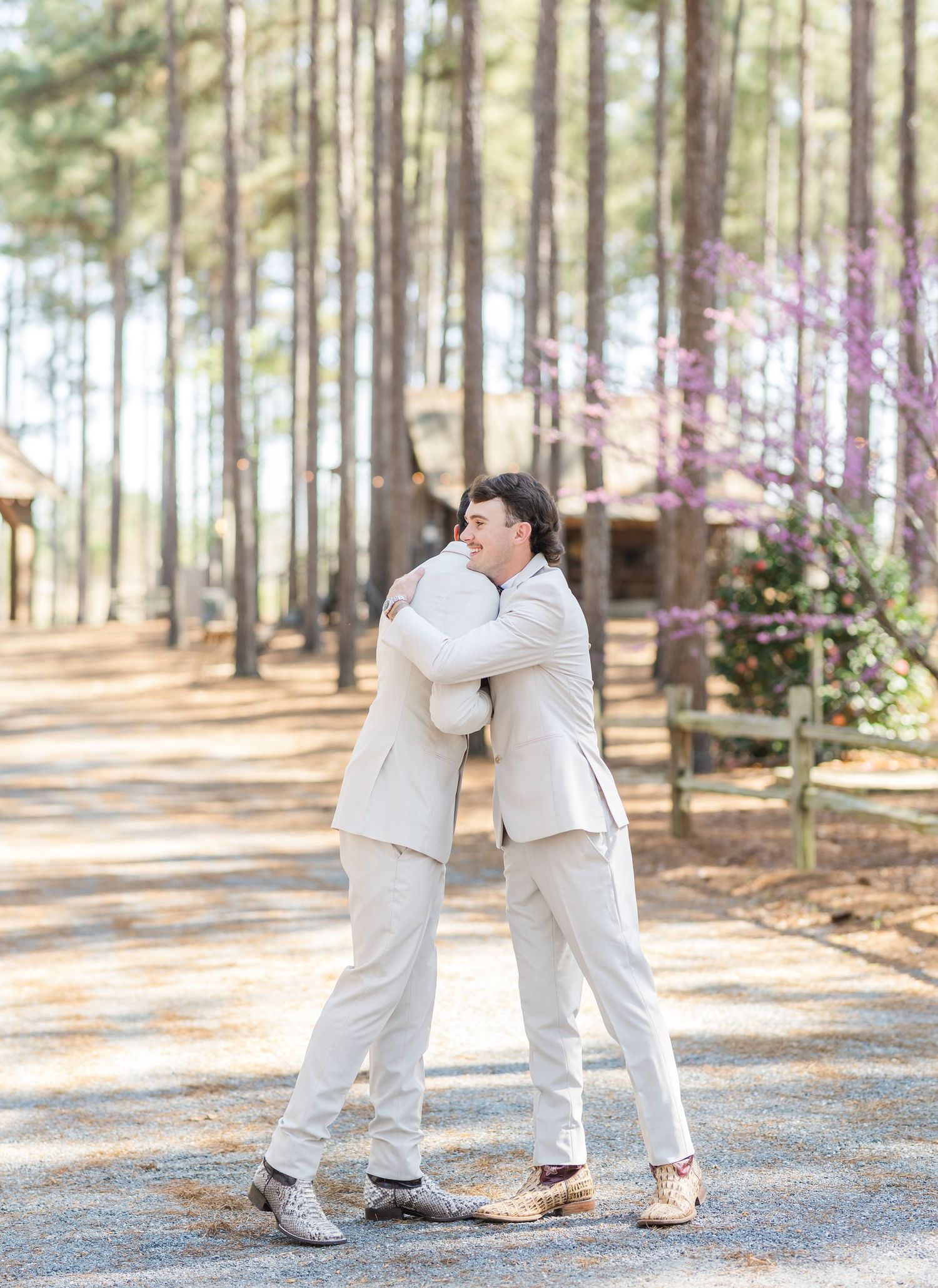 Two grooms in matching cream suits share a heartfelt embrace on a gravel path surrounded by tall pine trees.