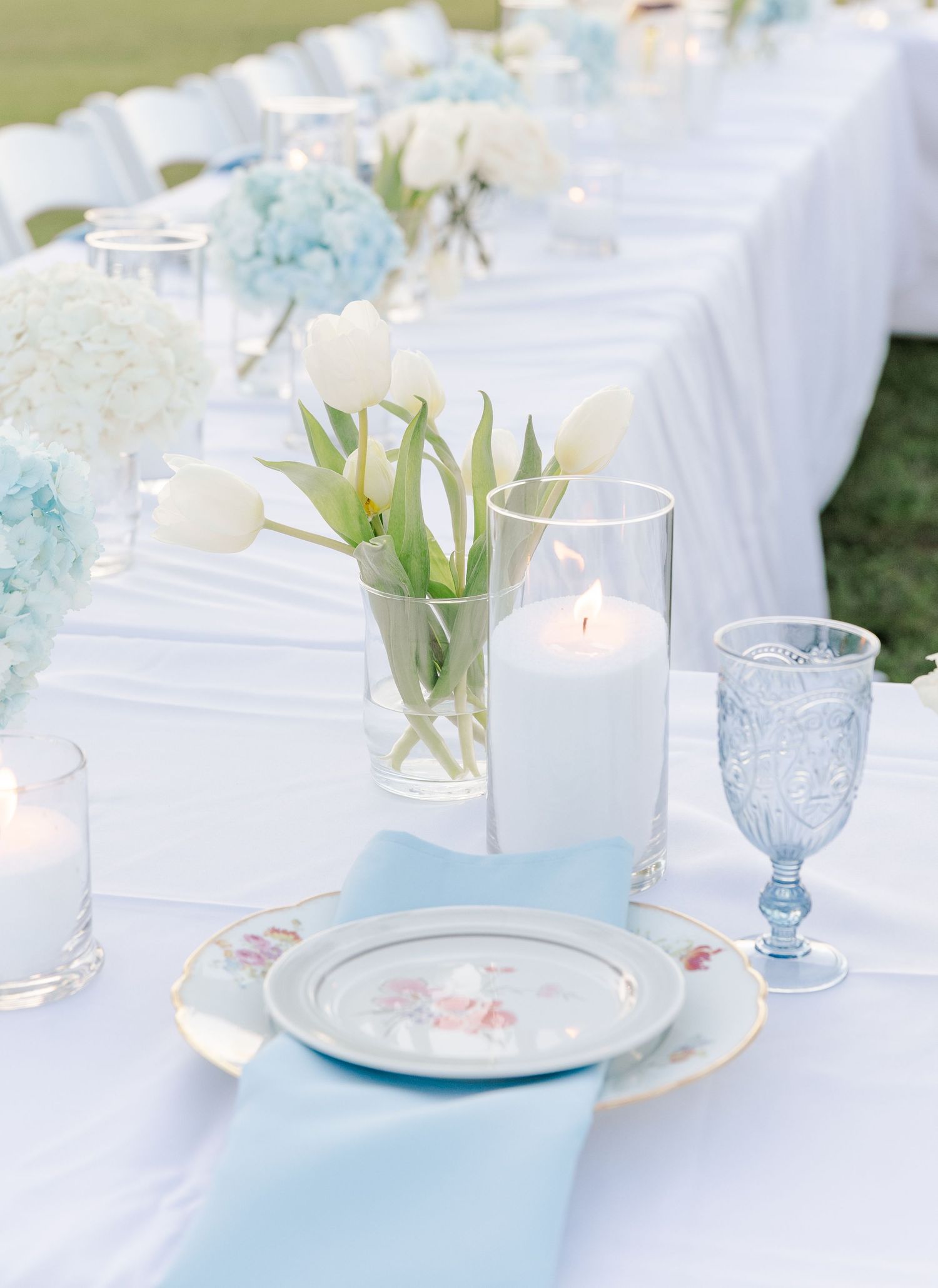 White wedding tablescape with blue hydrangeas, white tulips, candles, and blue glassware.