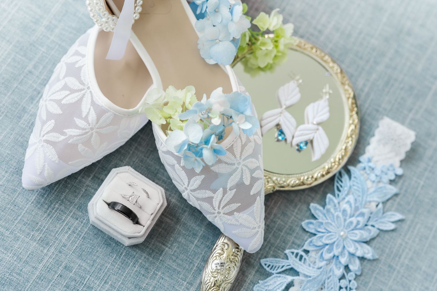 White lace bridal heels with blue hydrangea earrings, wedding rings, and a blue lace garter on a soft blue backdrop.