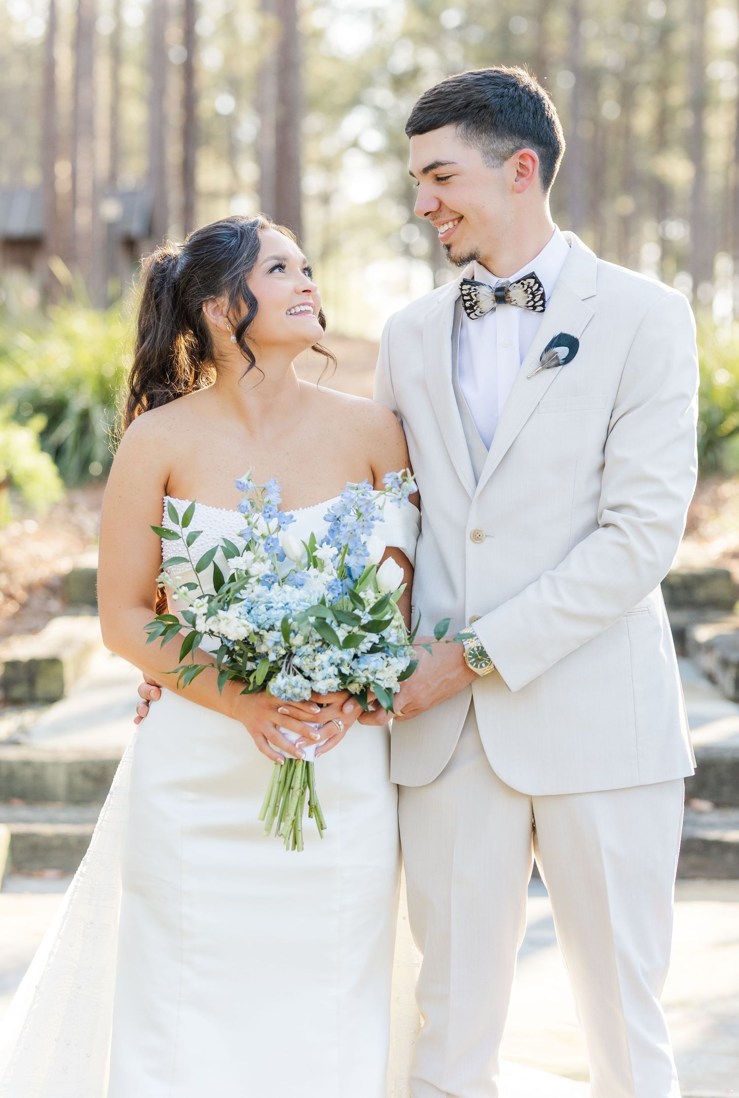Bride and groom smiling at each other outdoors, groom in cream suit with bow tie, bride holding blue bouquet.