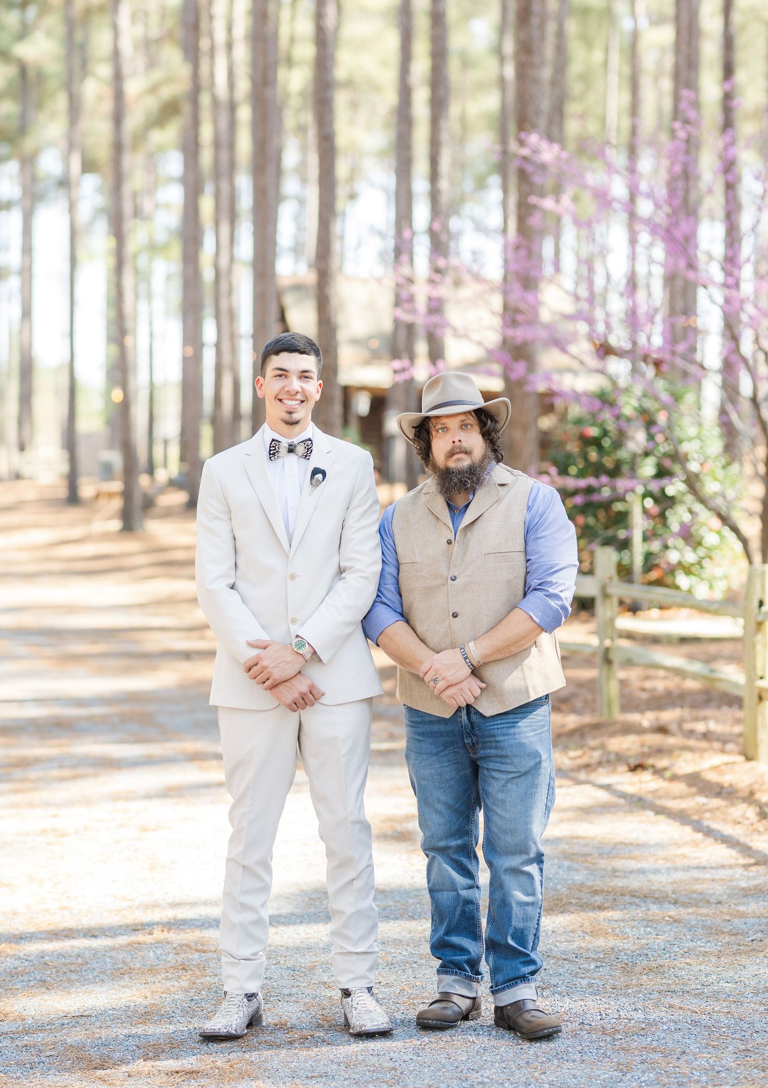 Groom in cream suit standing with man in cowboy hat and jeans at outdoor wedding.