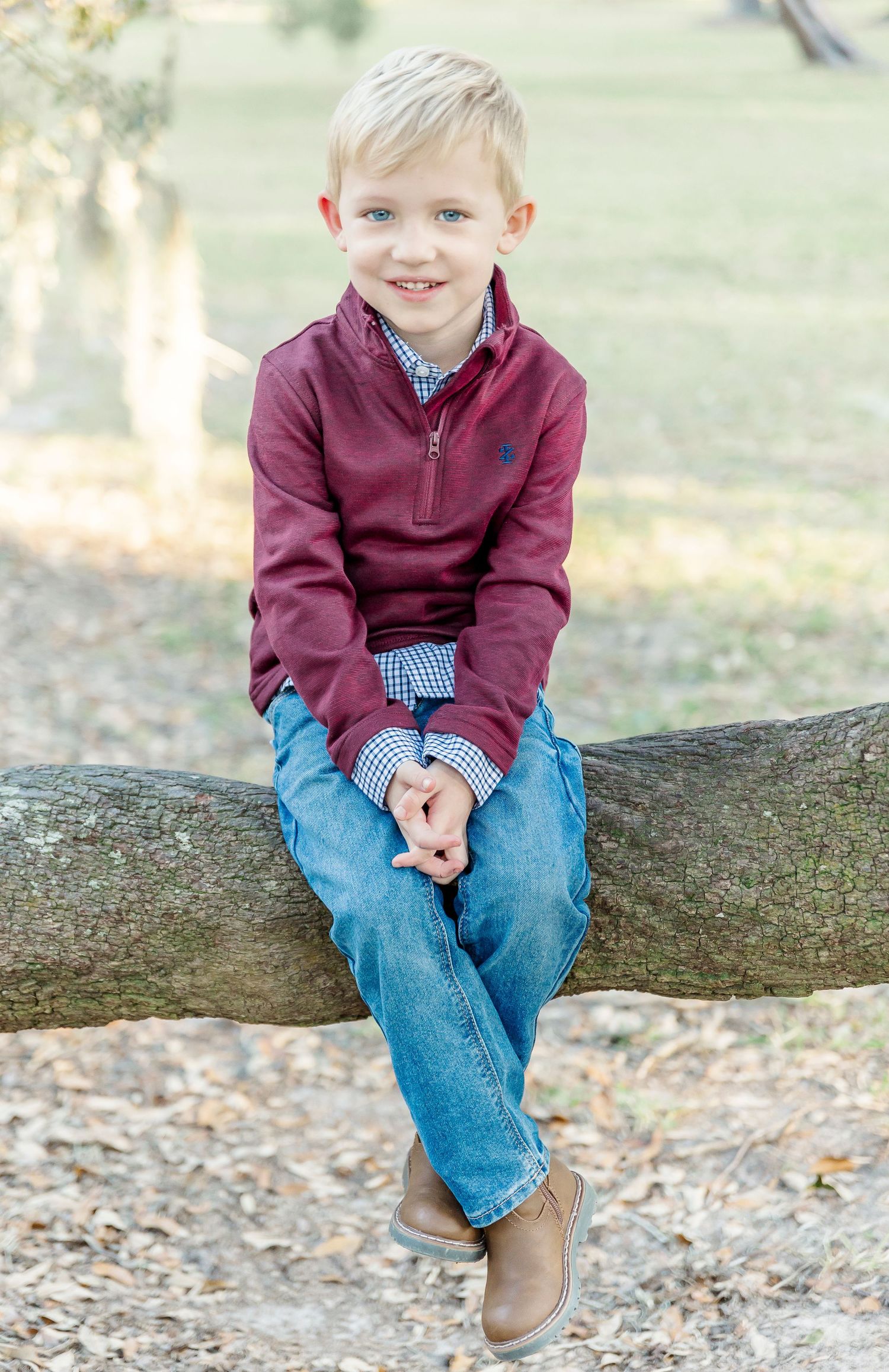 A child in burgundy pullover and jeans sits casually on a tree branch smiling at the camera.