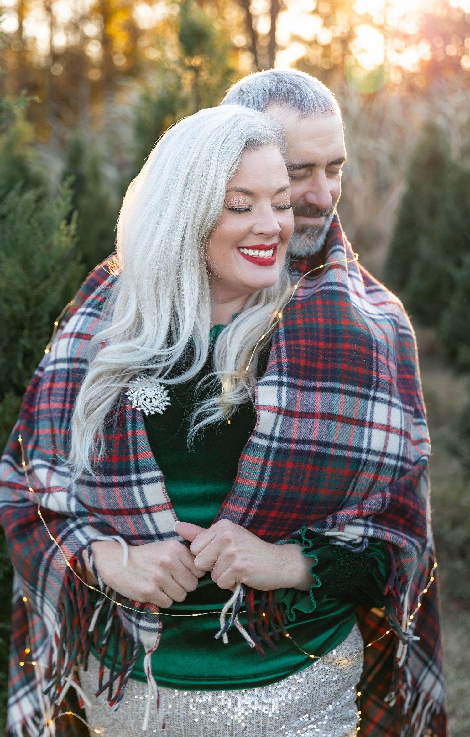 Couple wrapped in plaid blanket share intimate moment among evergreen trees at golden hour.