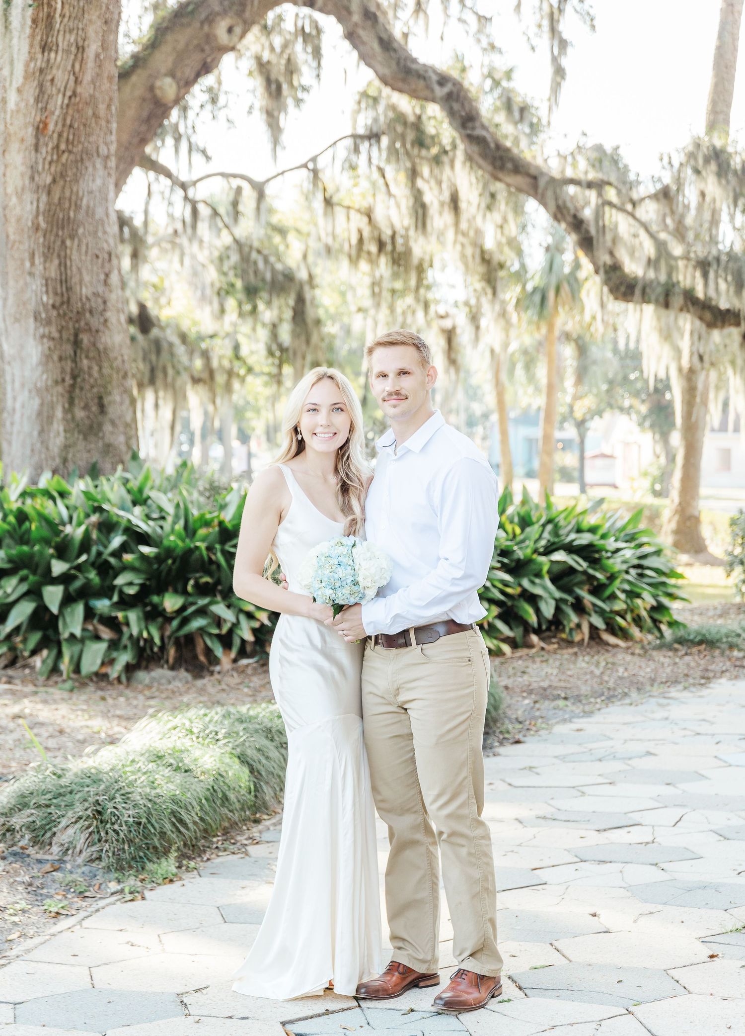 A couple poses under Spanish moss draped trees in a formal outdoor garden setting.