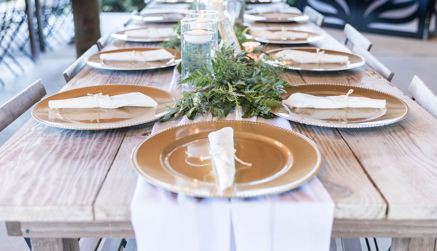 Elegant table setting with gold charger plates and greenery centerpiece on rustic wooden table.
