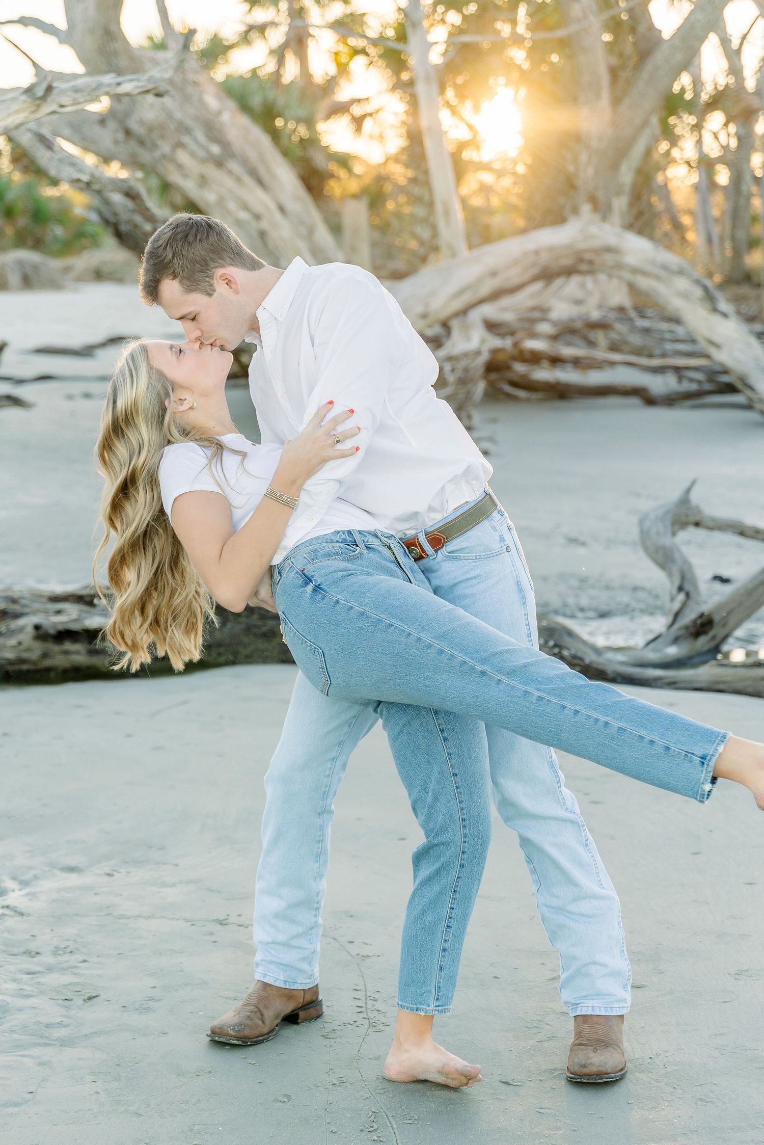 A romantic couple embraces in a dance-like pose on a beach with driftwood.