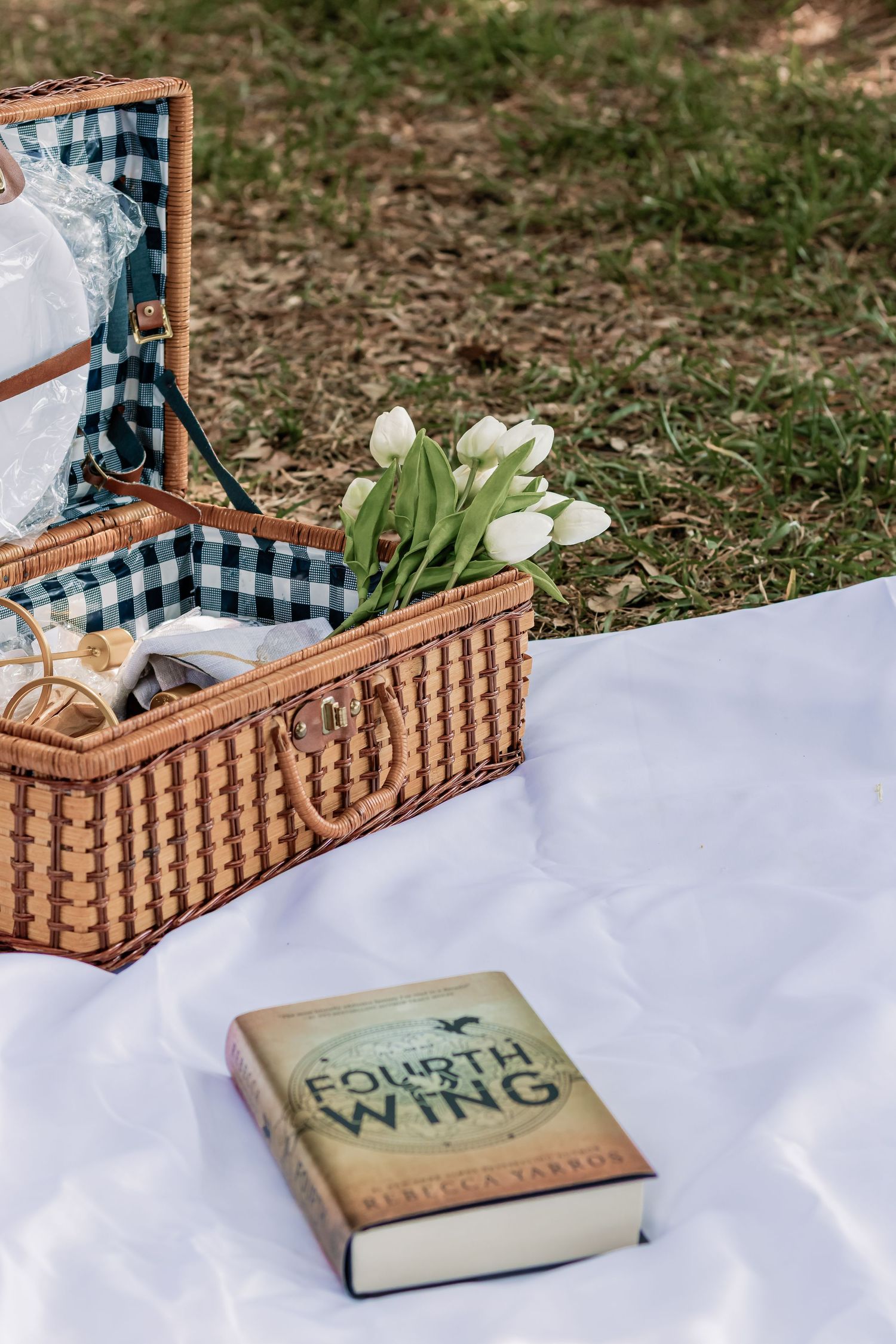 White tulips in picnic basket with book titled 'County Wine' on white blanket outdoors.