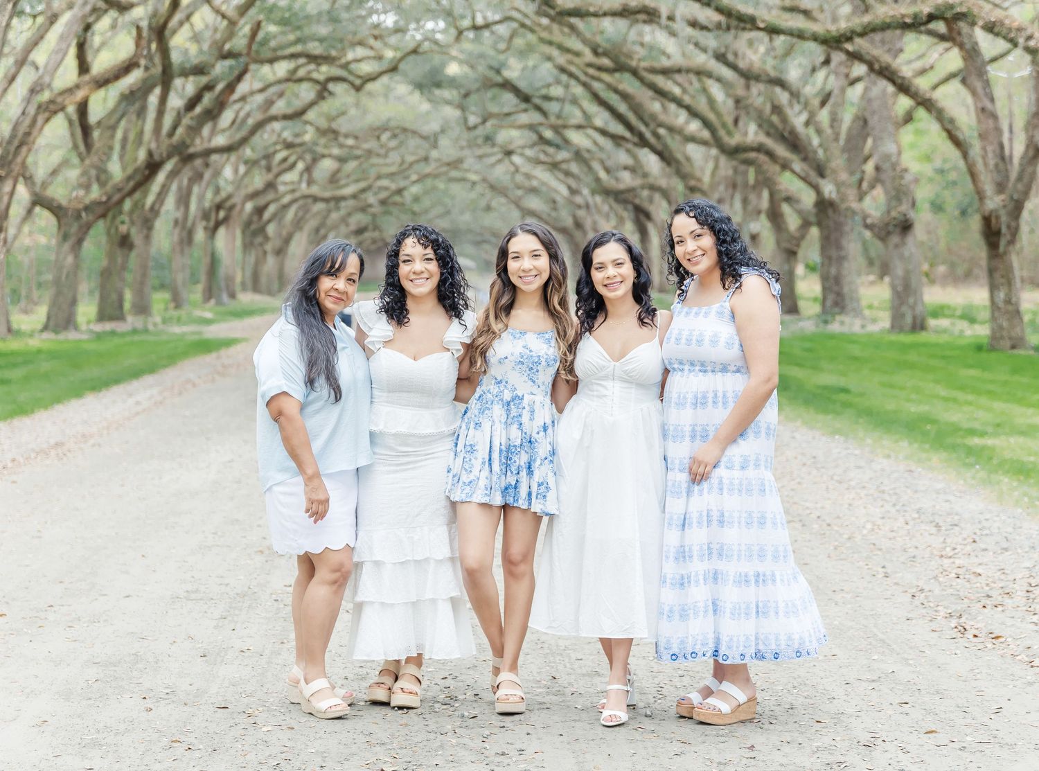 Five women in summer dresses walking together under oak tree canopy.