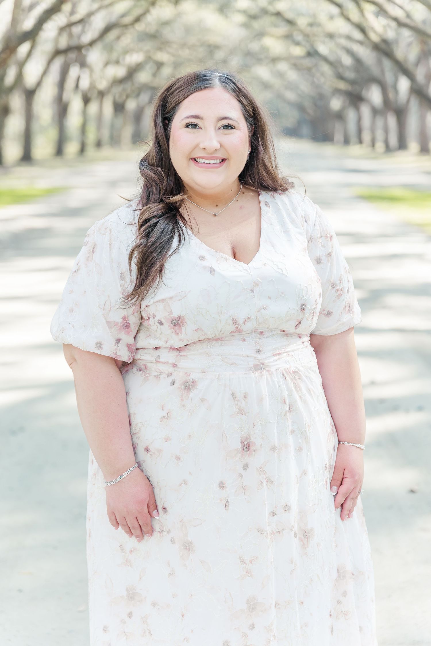 A beautiful woman in a floral white dress poses on a tree-lined path.