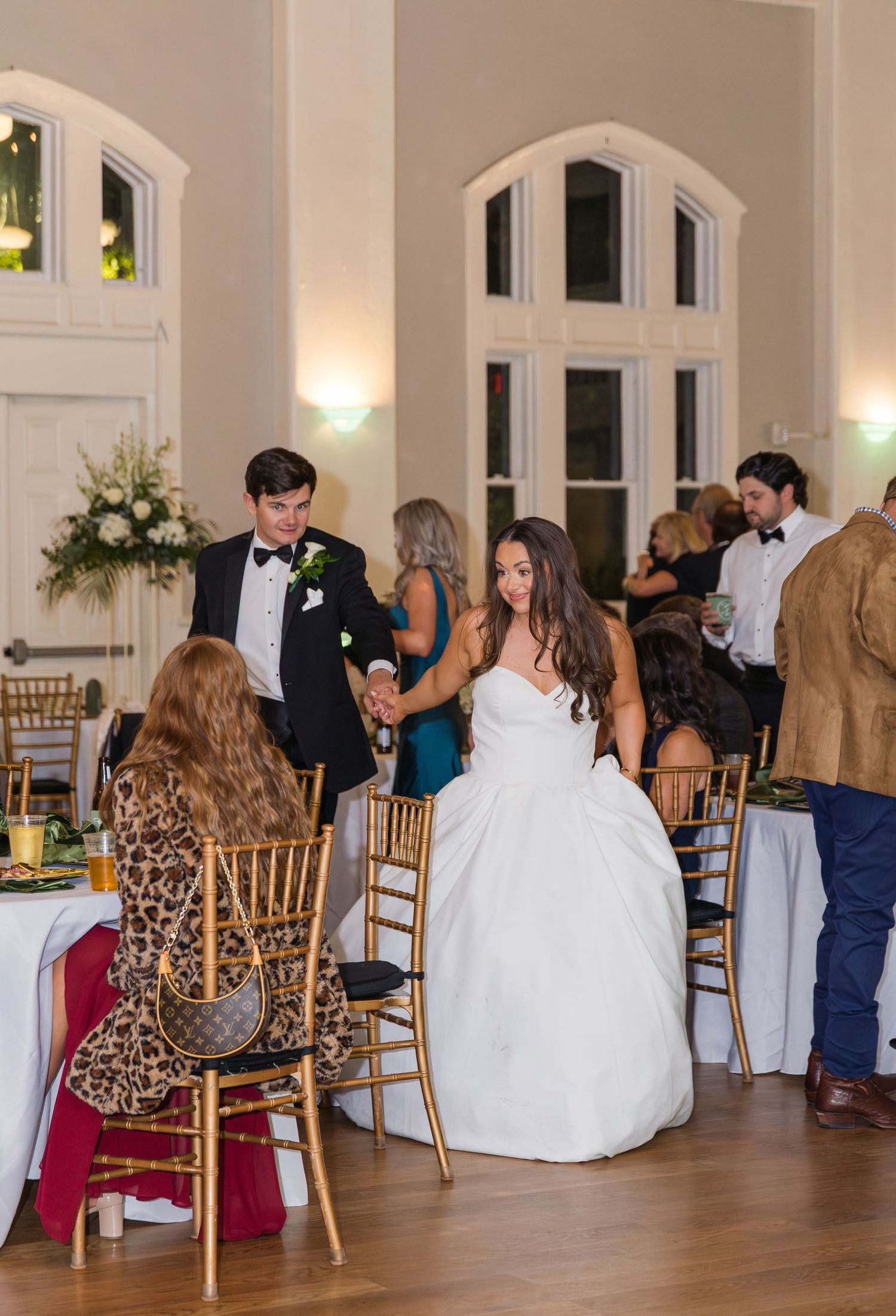 Bride in white ball gown and groom in black tuxedo greeting seated wedding guests at elegant reception venue.
