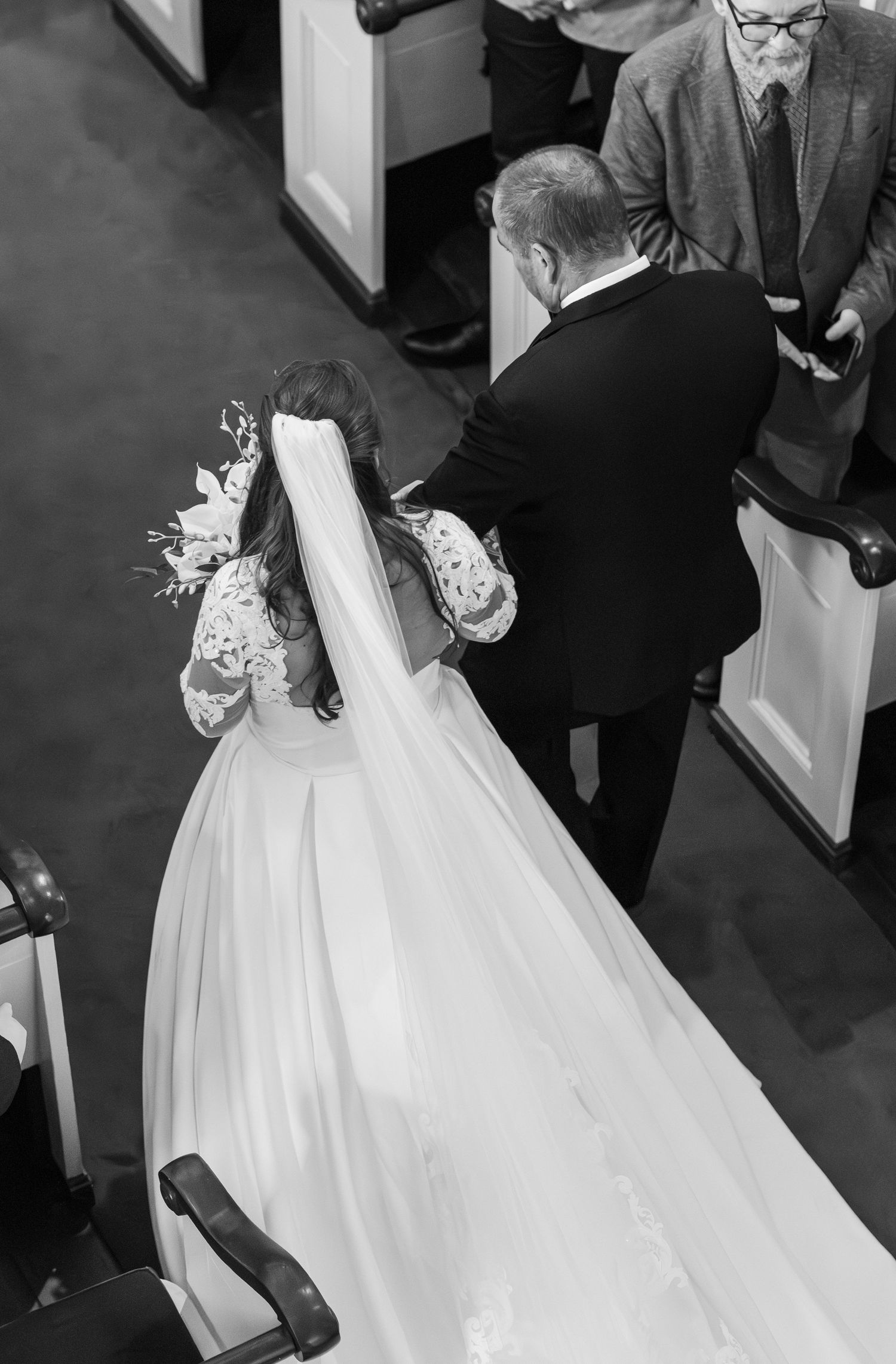 Black and white aerial photo of bride in lace gown and veil walking down church aisle with escort.