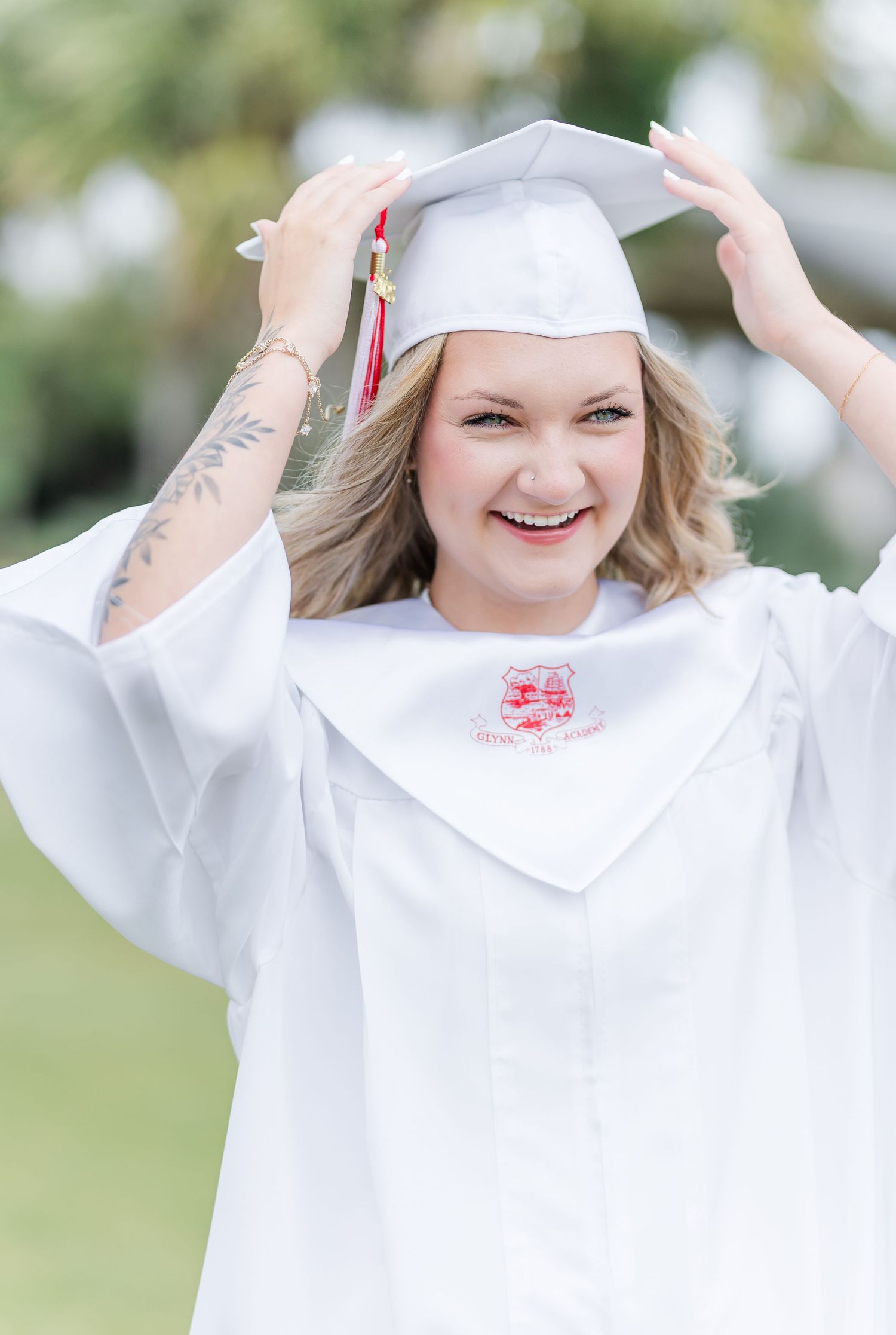 Joyful graduate in white cap and gown adjusts mortarboard while smiling outdoors.