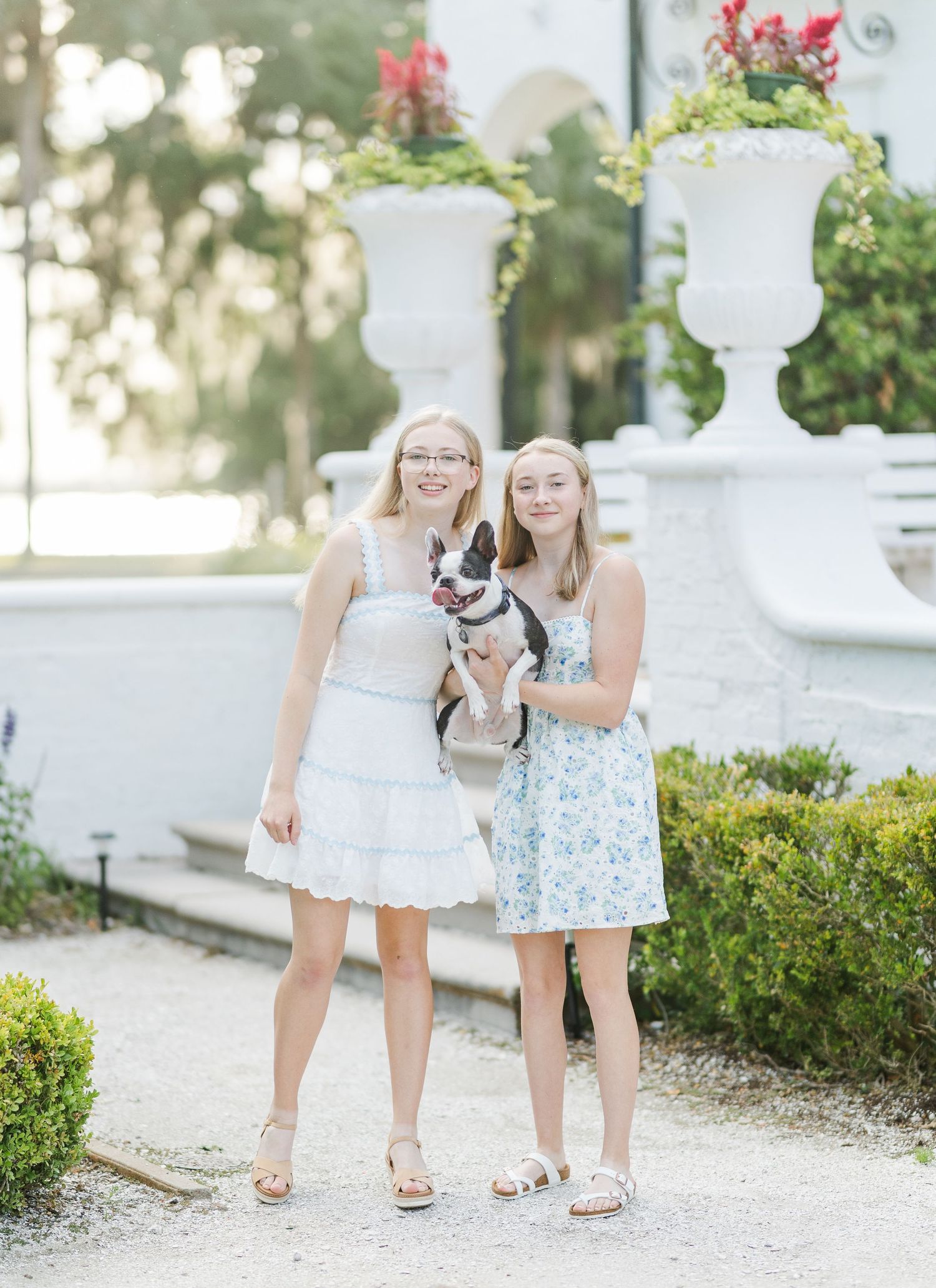 Two friends holding small dog pose together by decorative garden urns with red flowers.