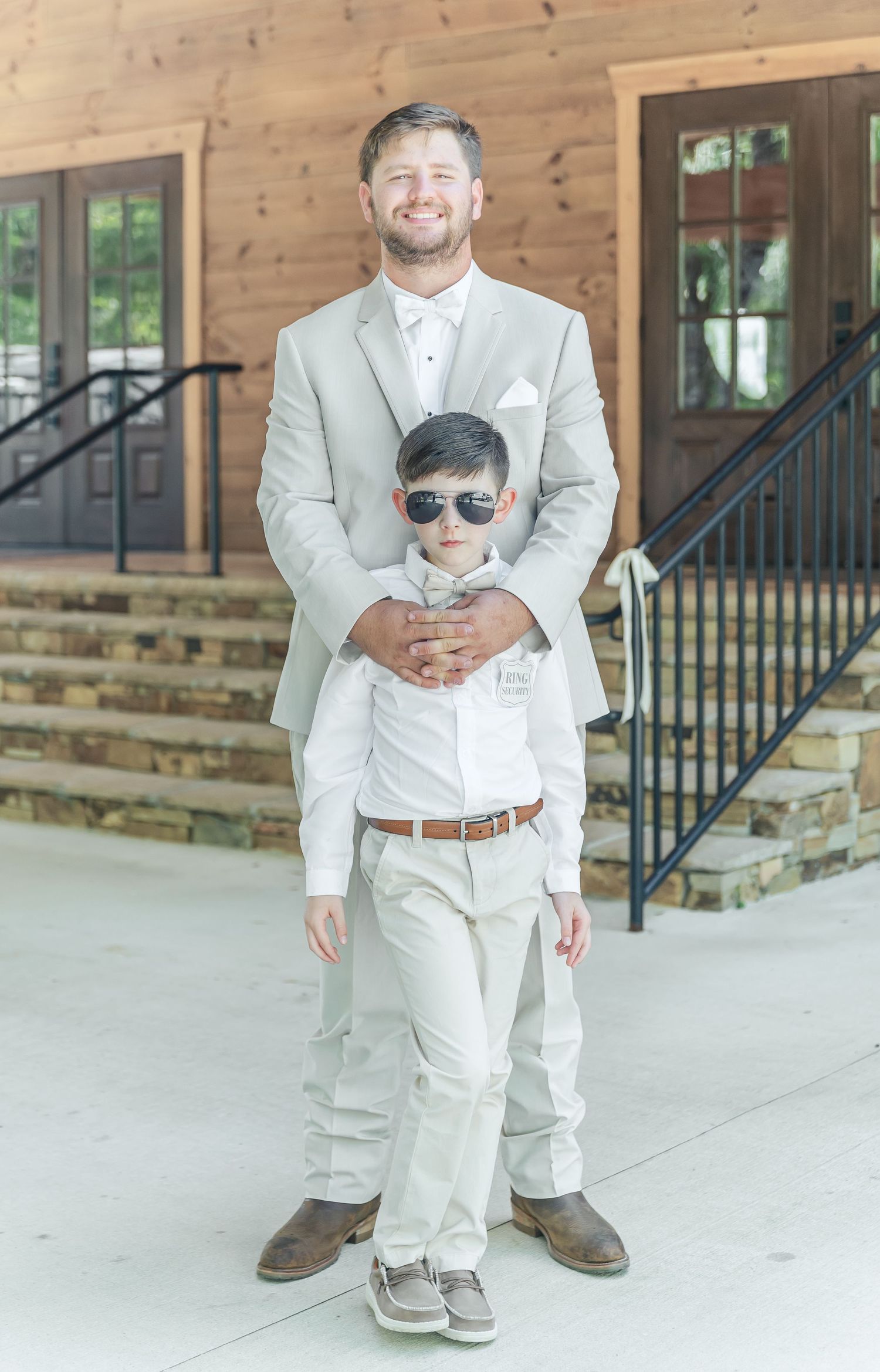 Person in light suit poses with young ring bearer wearing sunglasses on wooden porch.