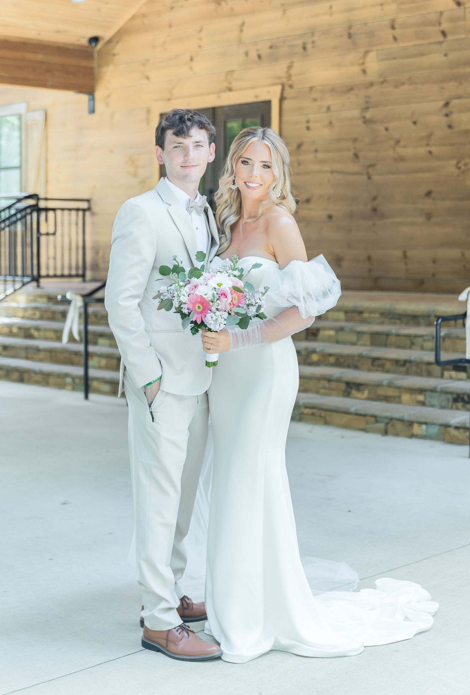 Wedding couple in matching white attire pose on rustic wooden porch with floral bouquet.