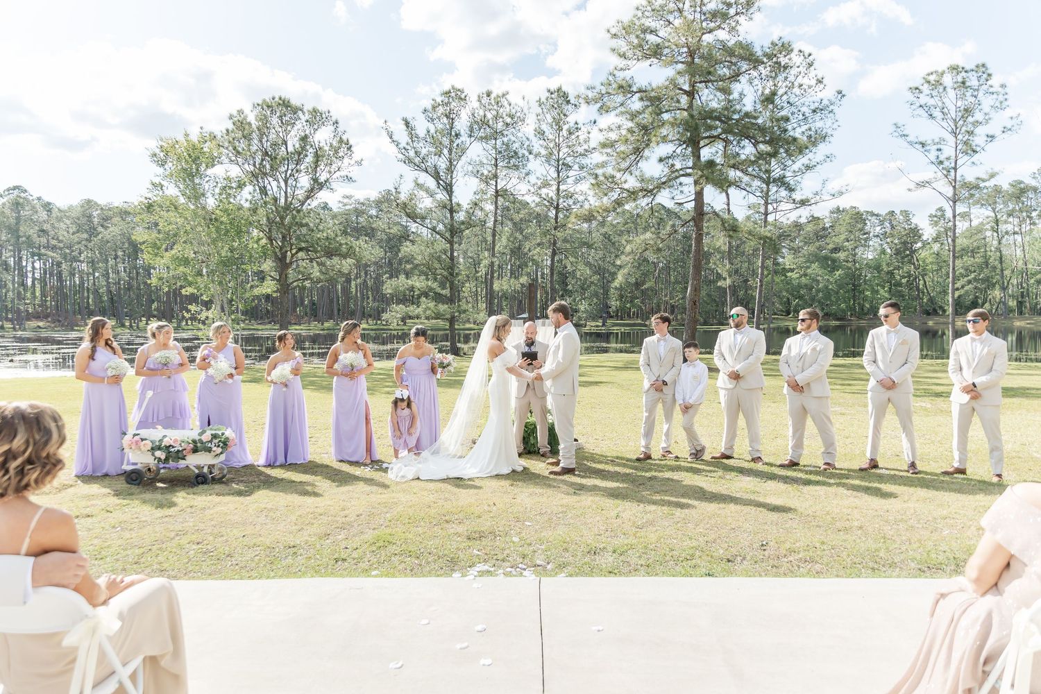 Wedding party lined up during a sunny outdoor ceremony with bridesmaids in lavender and groomsmen in light suits.