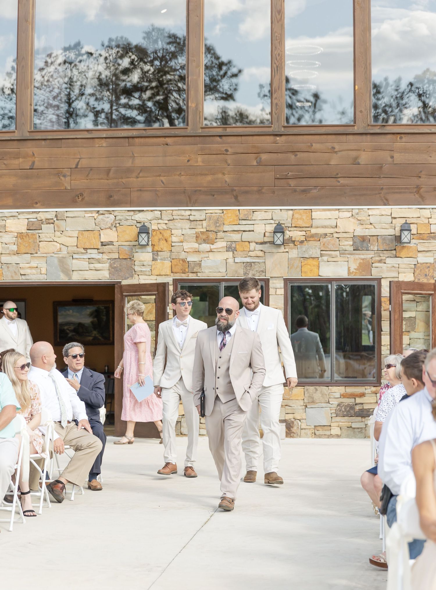 Wedding party members in light colored suits walk down the aisle at an outdoor ceremony venue.