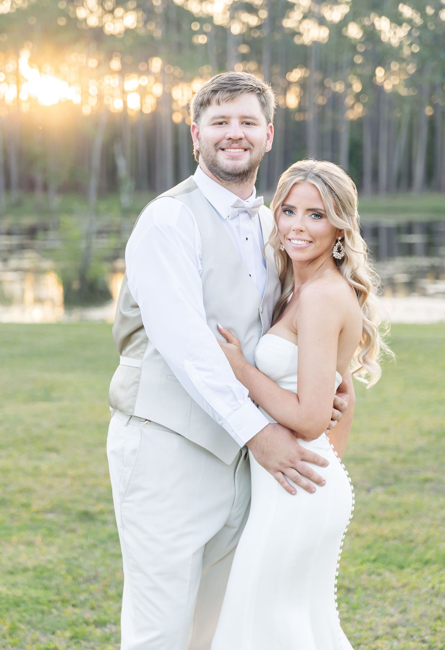 Sunset wedding portrait of newlyweds in light-colored attire by a serene lake with pine trees.