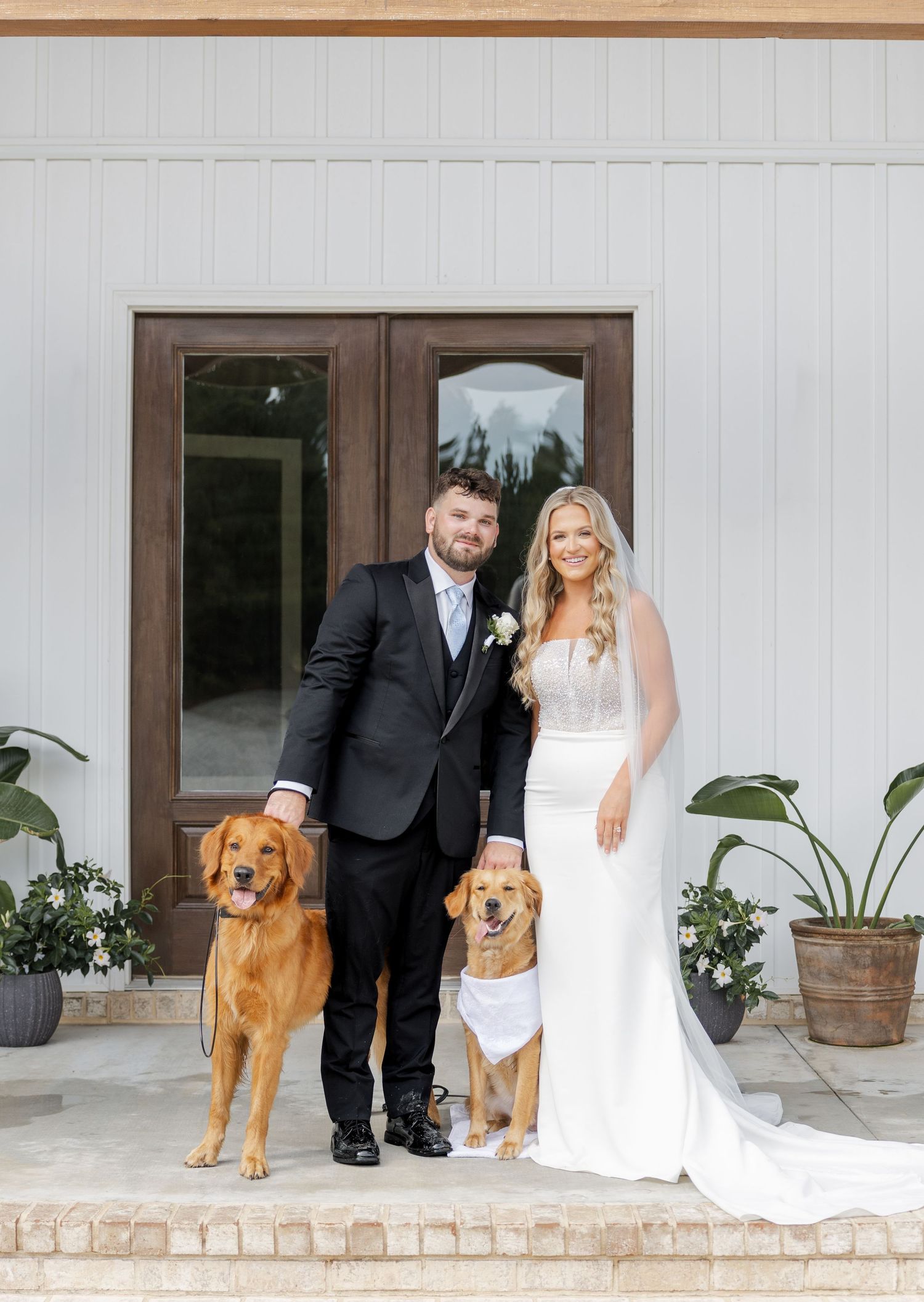 Newlyweds posing with two golden retrievers, one wearing white bandana, outside white barn wedding venue.