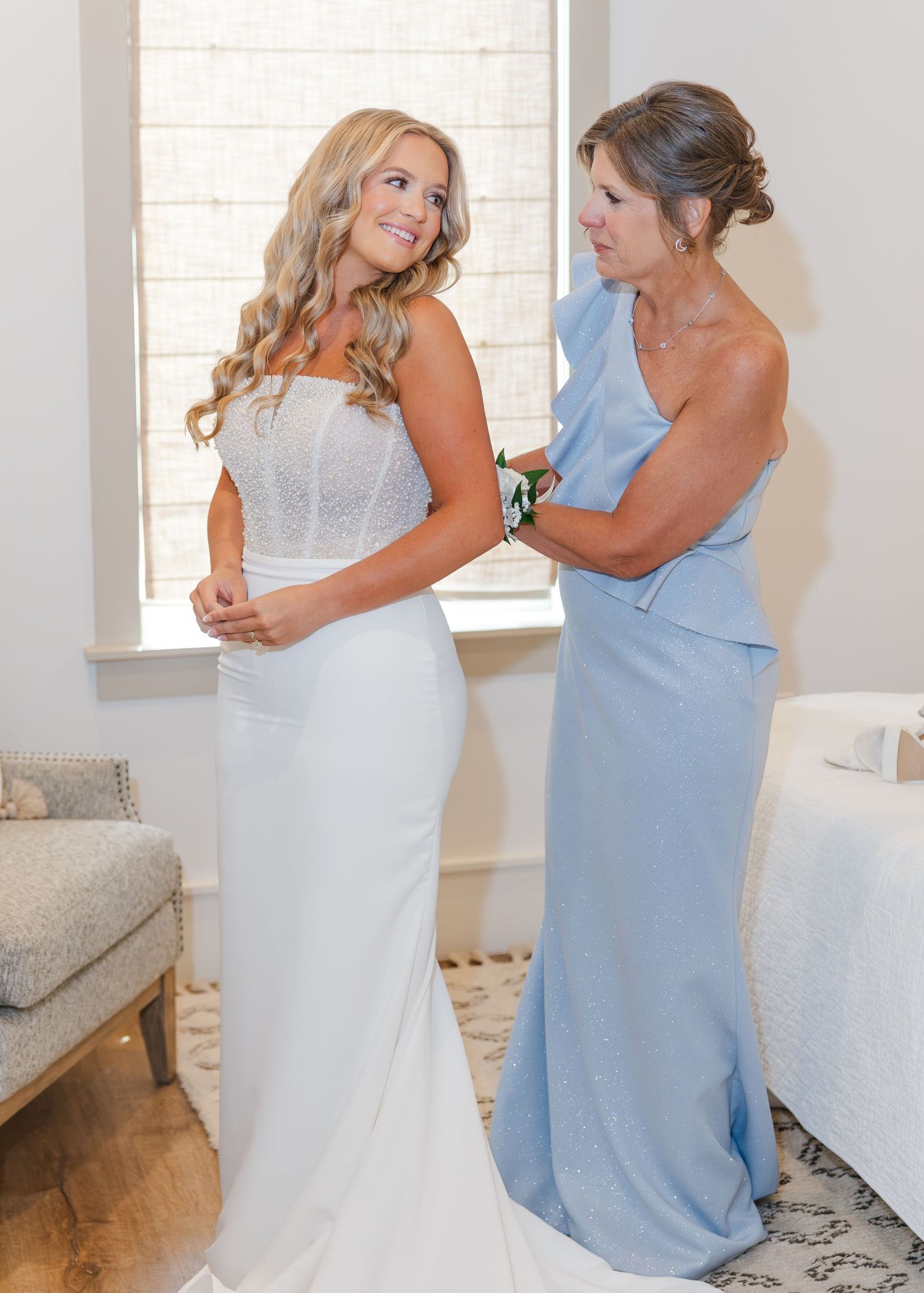 Bride in strapless white wedding gown being helped by mother in blue dress near a bright window.