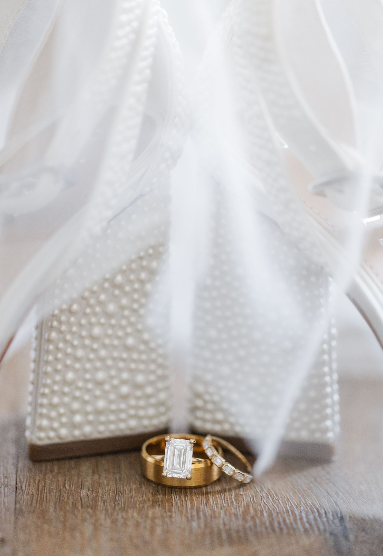 Gold wedding bands and emerald-cut diamond engagement ring displayed against pearl-studded bridal heels and white veil.